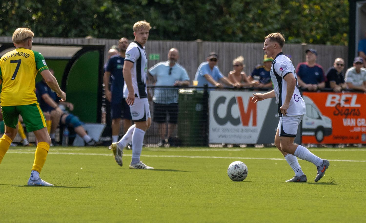 Two footballers in black and white kits communicate on the pitch during a sunny match against a team in yellow and green.