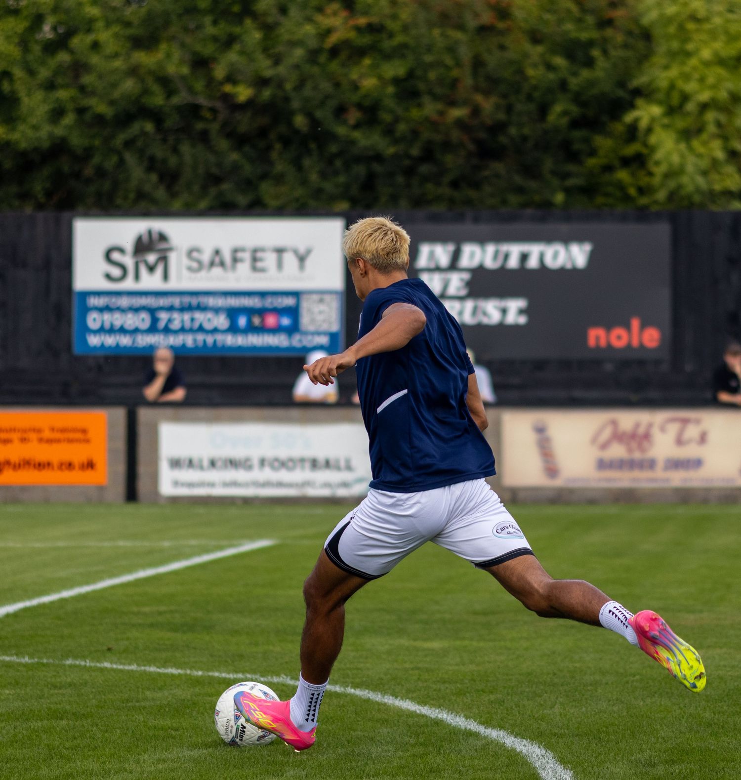 Blonde footballer in navy kit striking a ball during pre-match warmup with SM Safety advertising boards behind.