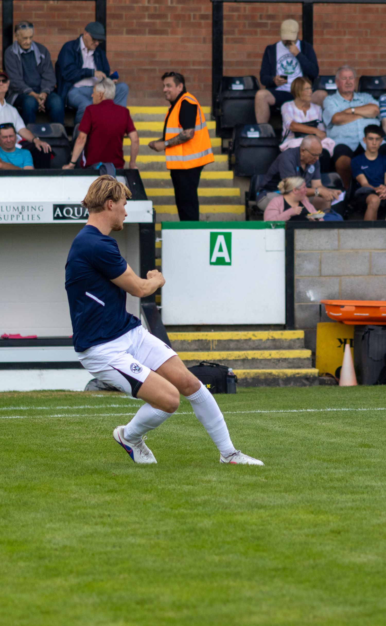 A footballer in navy blue kit celebrates a goal on the pitch with spectators in the stands behind.
