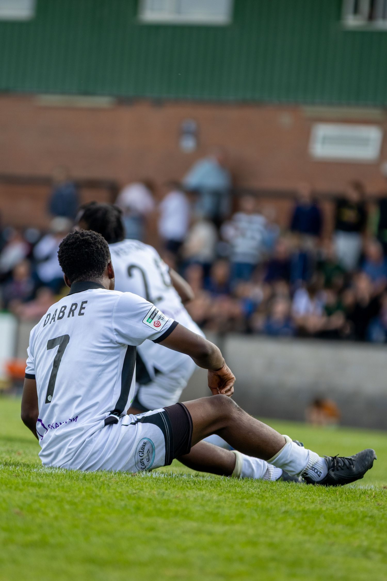 Football player wearing number 7 DABRE jersey sits on grass pitch during a soccer match with spectators in background.