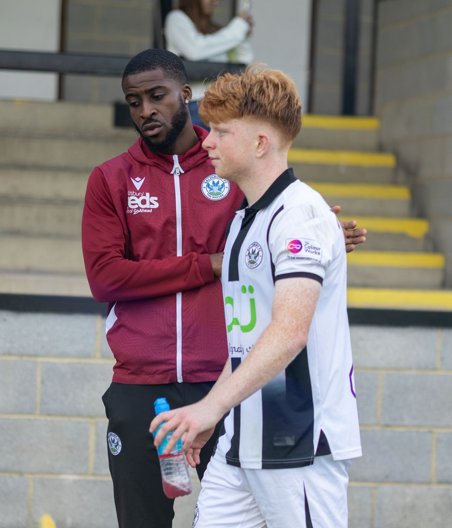 Two football players, one in maroon tracksuit and one in black and white kit, having a conversation on the sidelines.