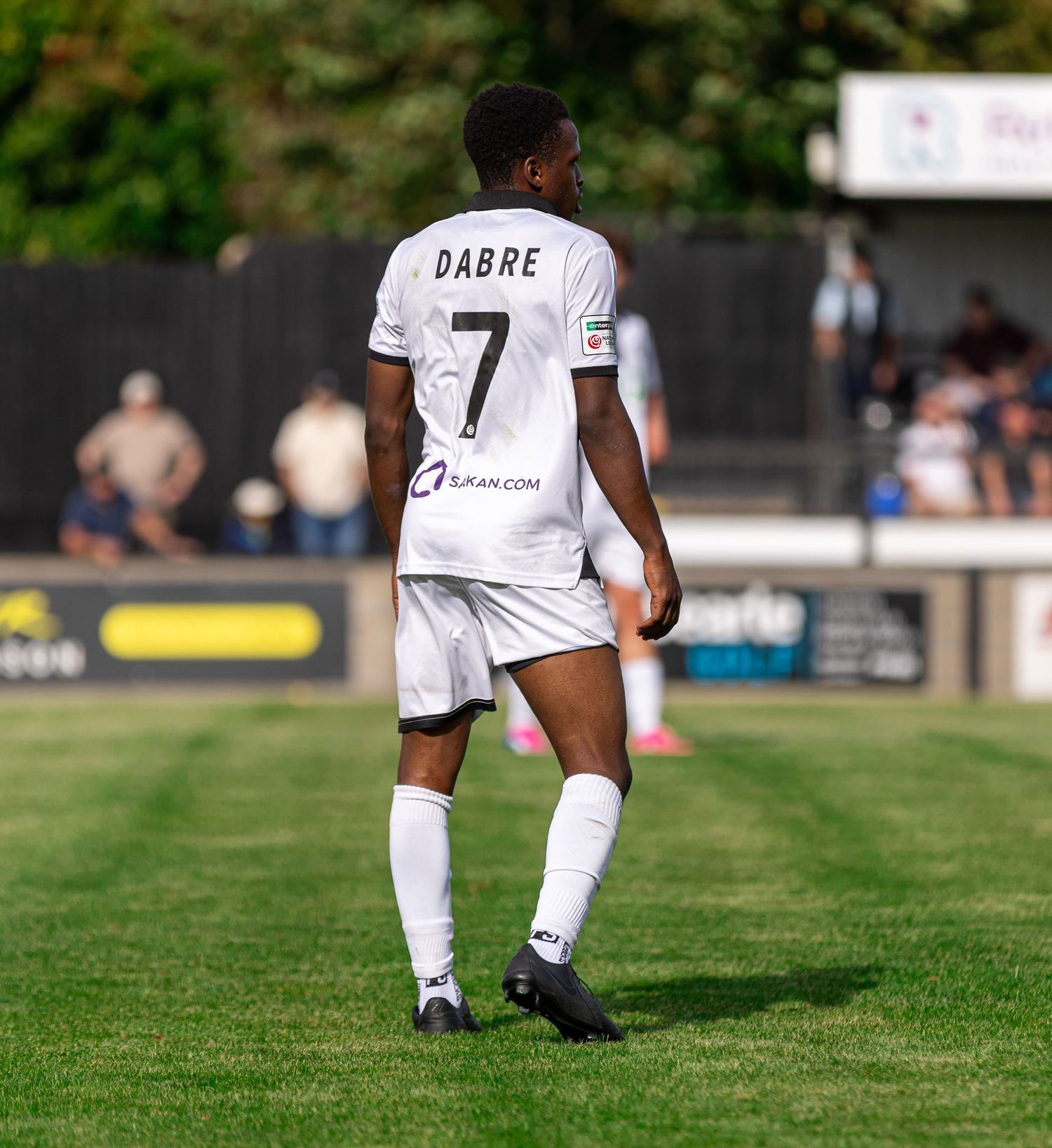 Soccer player wearing white jersey with number 7 and name DABRE stands on green pitch during a sunny match.