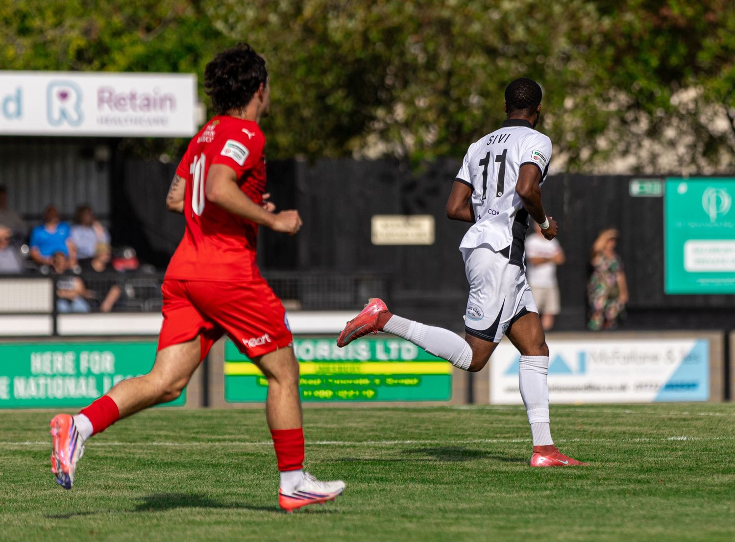 Two soccer players in red and white uniforms compete on a green pitch during a sunny outdoor match.
