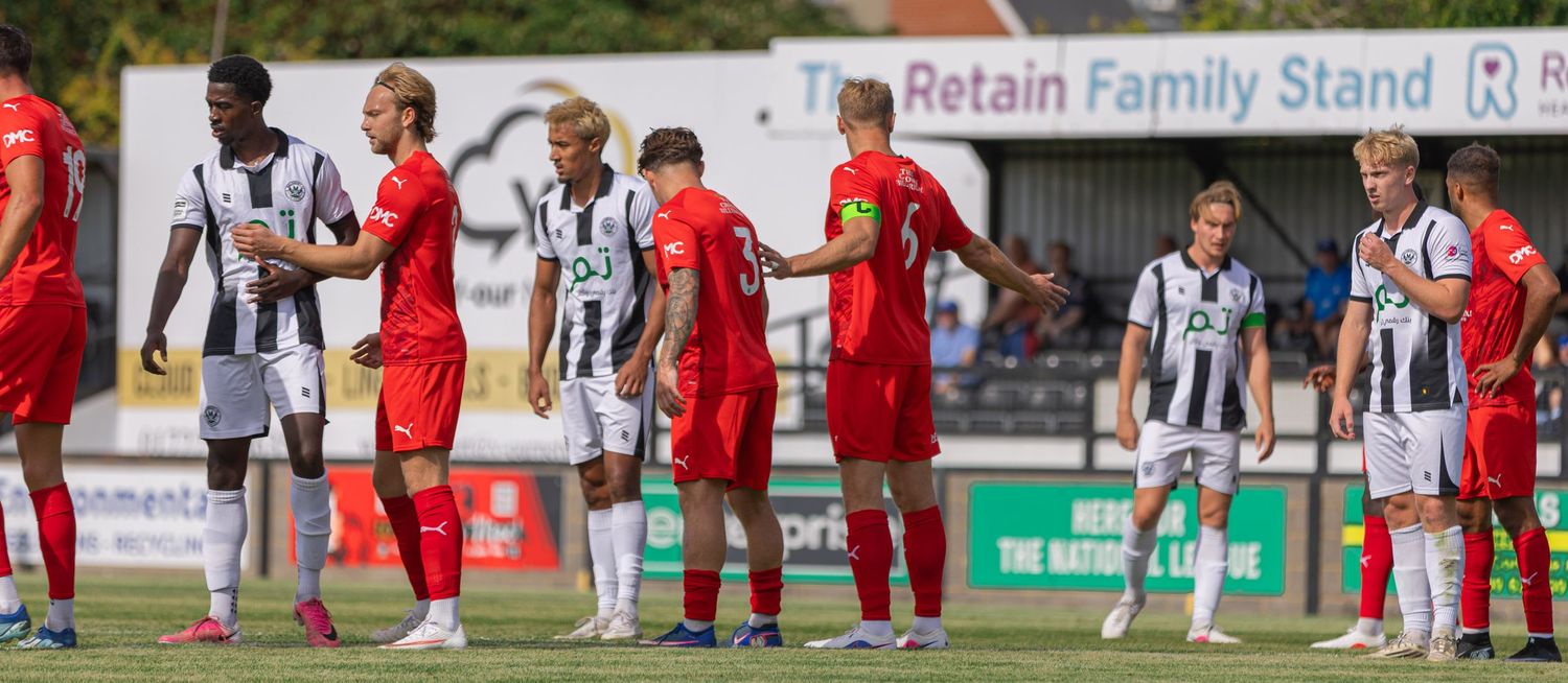 Football players in black-and-white and red kits line up on a sunny pitch near the Retain Family Stand.