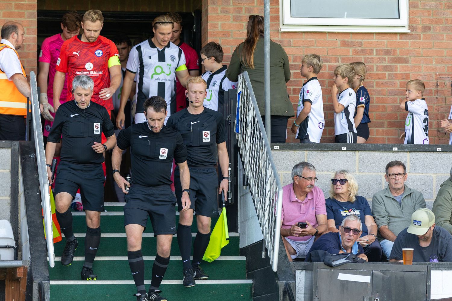 Football referees lead players onto the pitch at a sunny non-league match, with spectators watching from the stands.