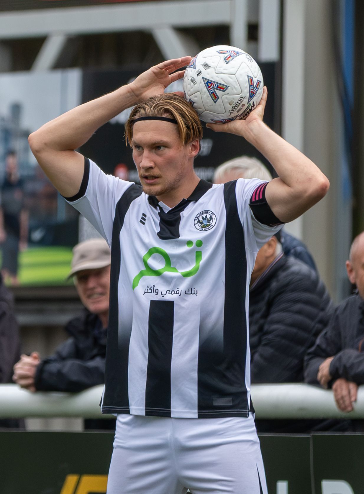 Football player in black and white striped jersey preparing to take a throw-in, holding ball above head on sideline.