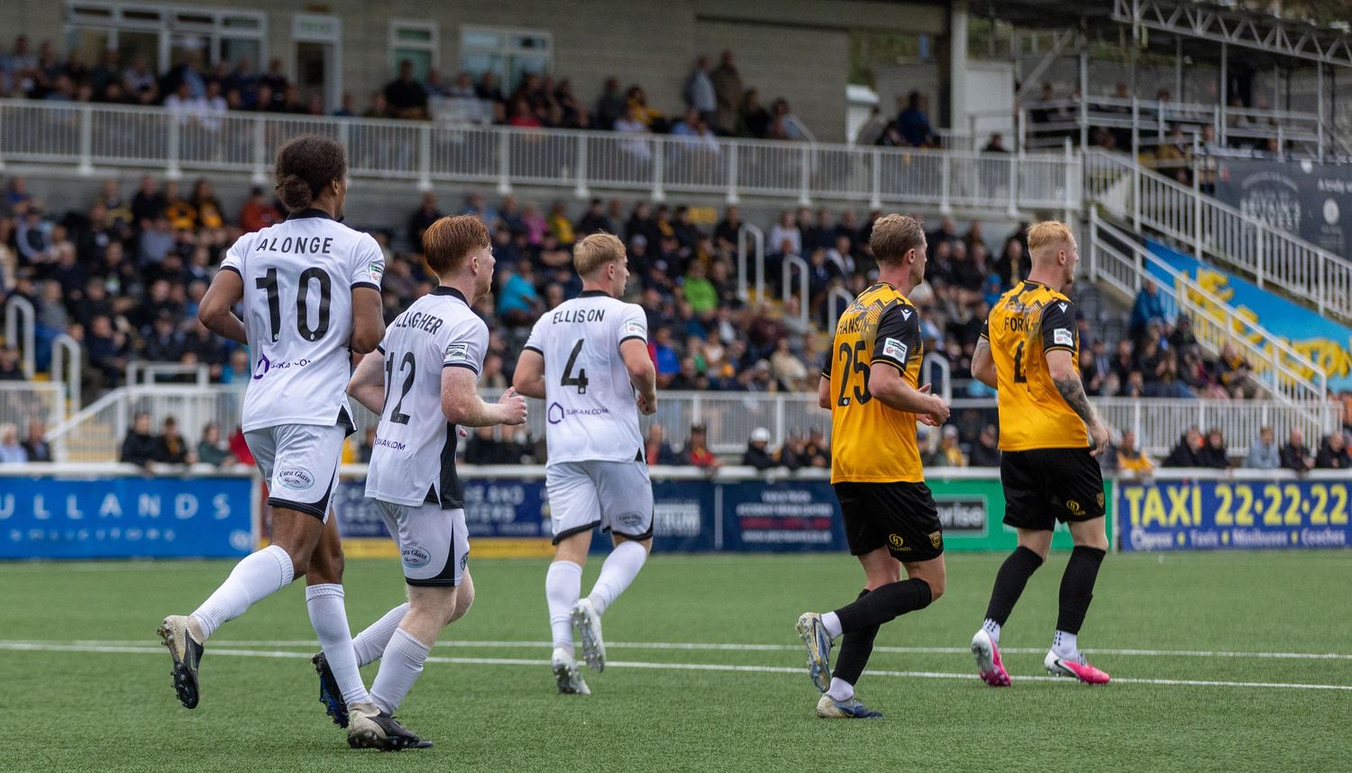 Soccer players in white celebrate a goal during a match against opponents in yellow and black on a sunny day.