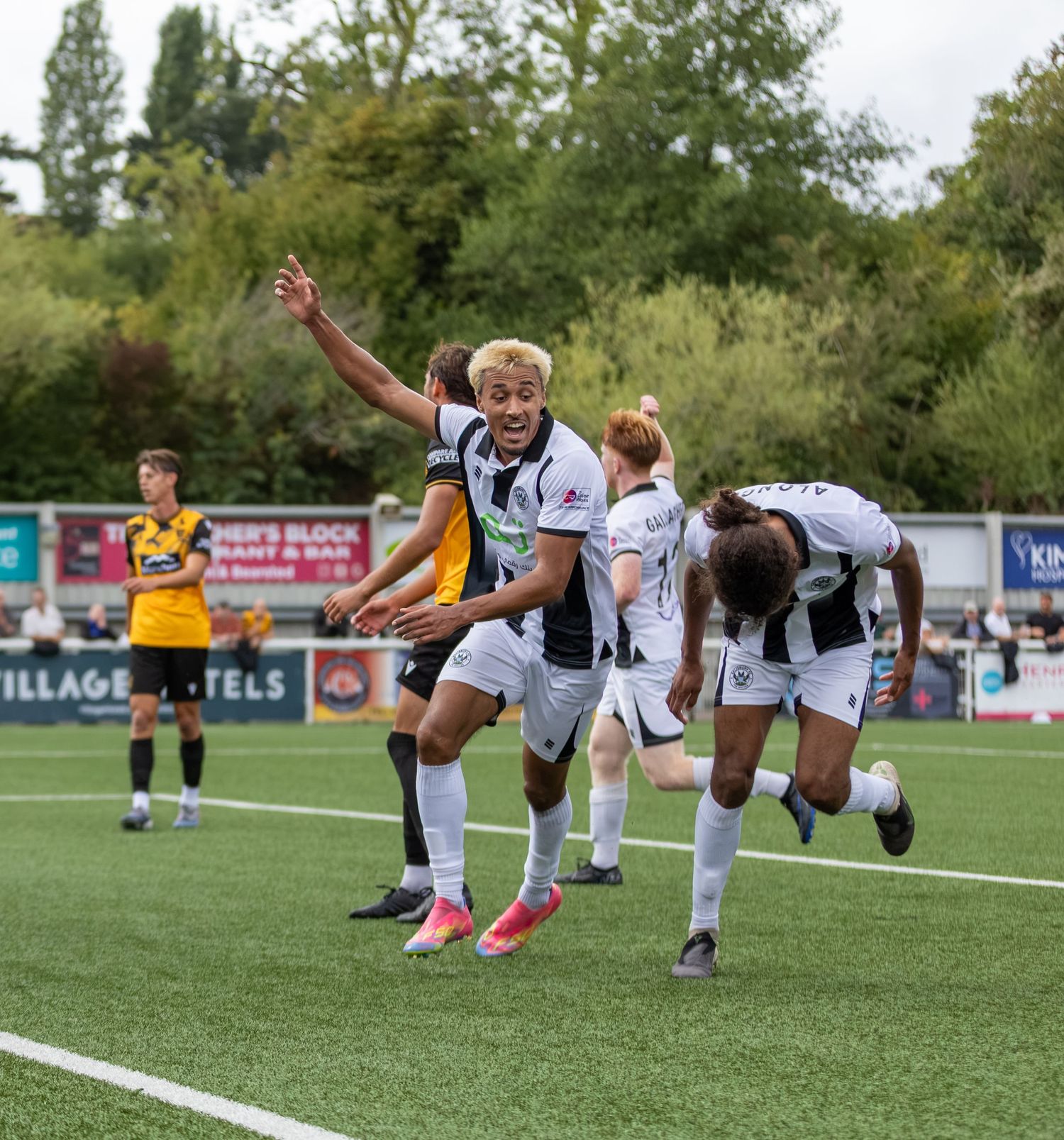 Soccer players in black and white uniforms celebrate a goal on a green pitch during a sunny outdoor match.