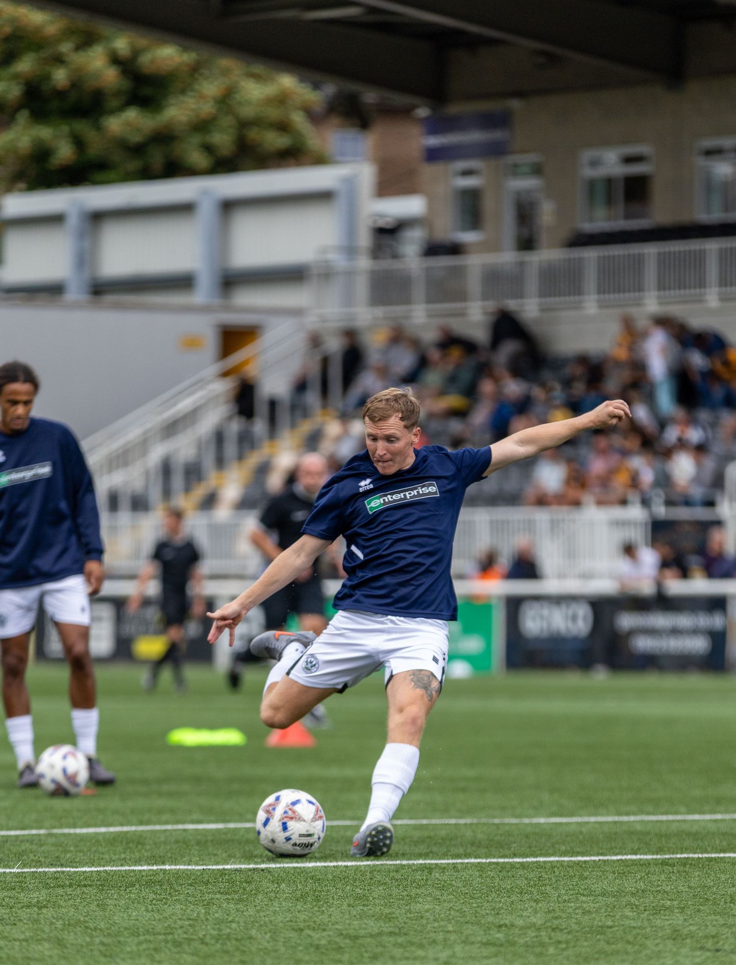 Soccer player in navy kit strikes ball during warm-up on green pitch with stadium stands in background.