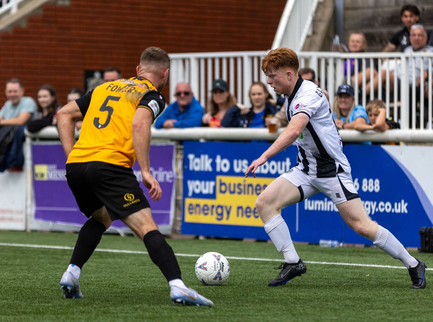 Two soccer players compete for the ball during a match, one wearing yellow and black, the other in white, on a sunny day.