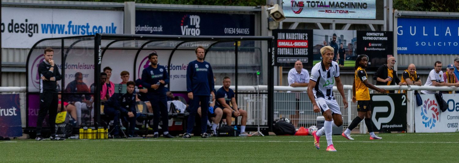 A football match in progress at a non-league ground with players on the pitch and coaching staff watching from the dugout.
