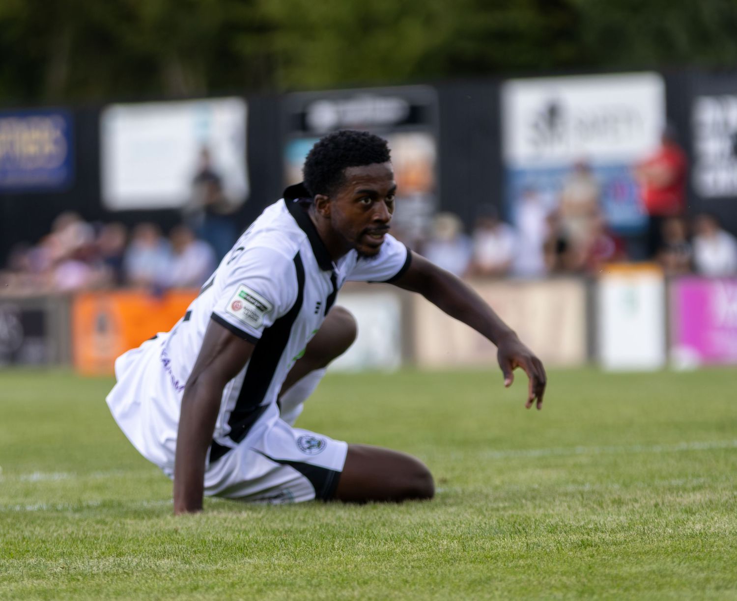 Non-league footballer in white and black kit sits on grass pitch after a challenge during a match.