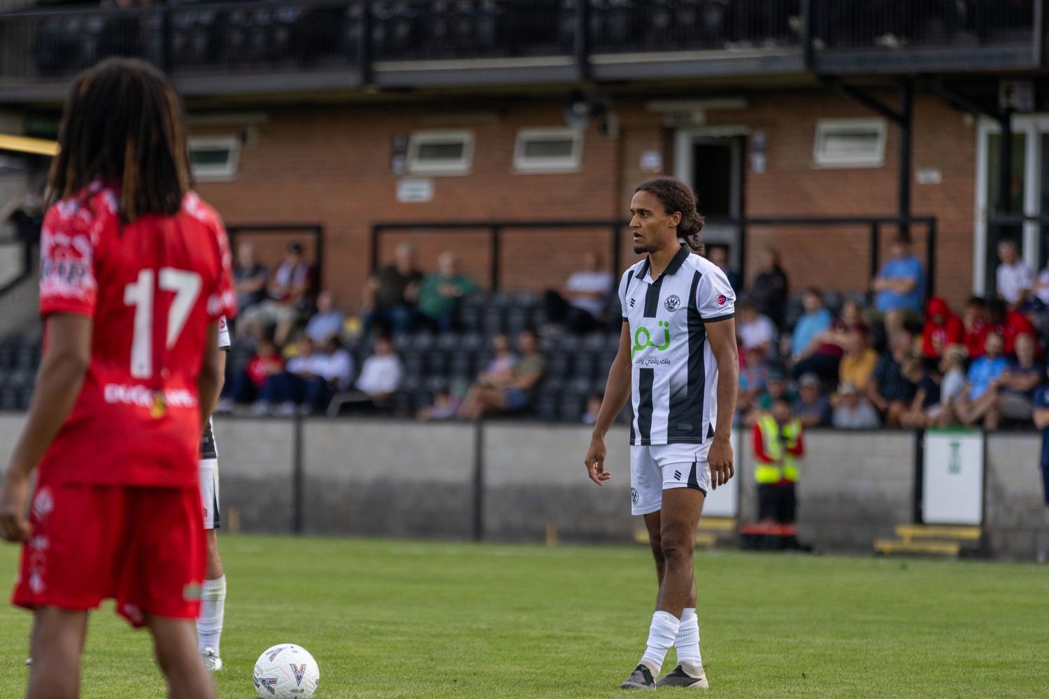 Female footballer in black and white kit prepares for free kick against red opponents.