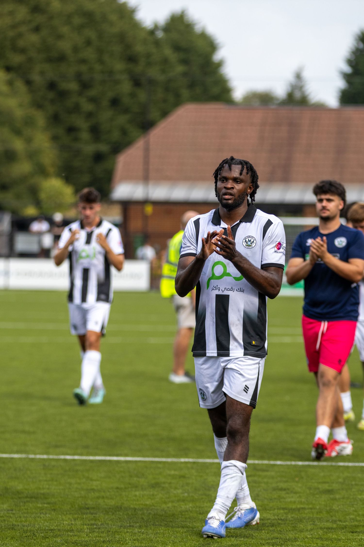 A footballer with dreadlocks in black and white striped kit claps fans after a match on a green pitch.