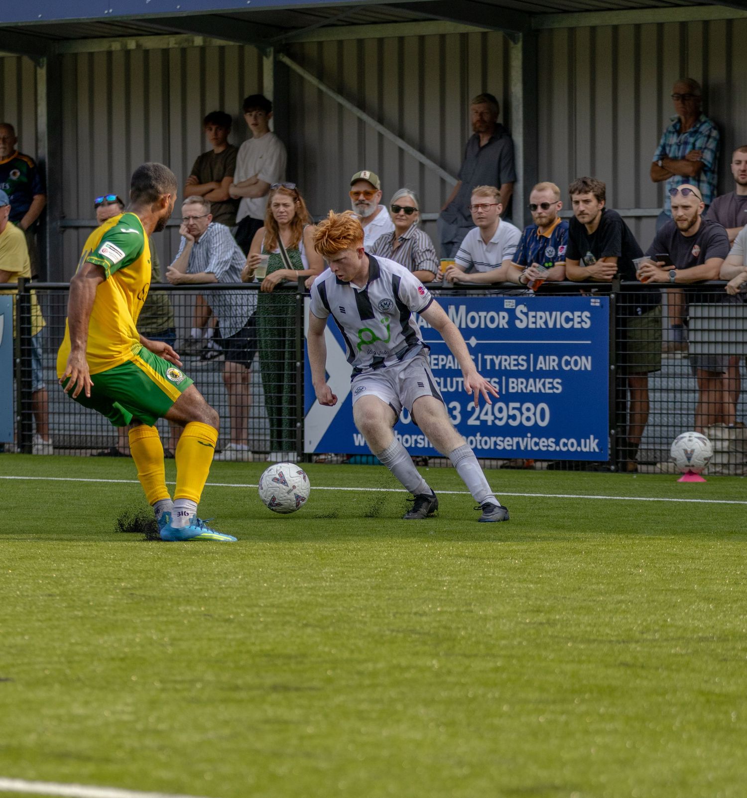Football player in white kit shields ball from opponent in yellow-green kit during competitive match.