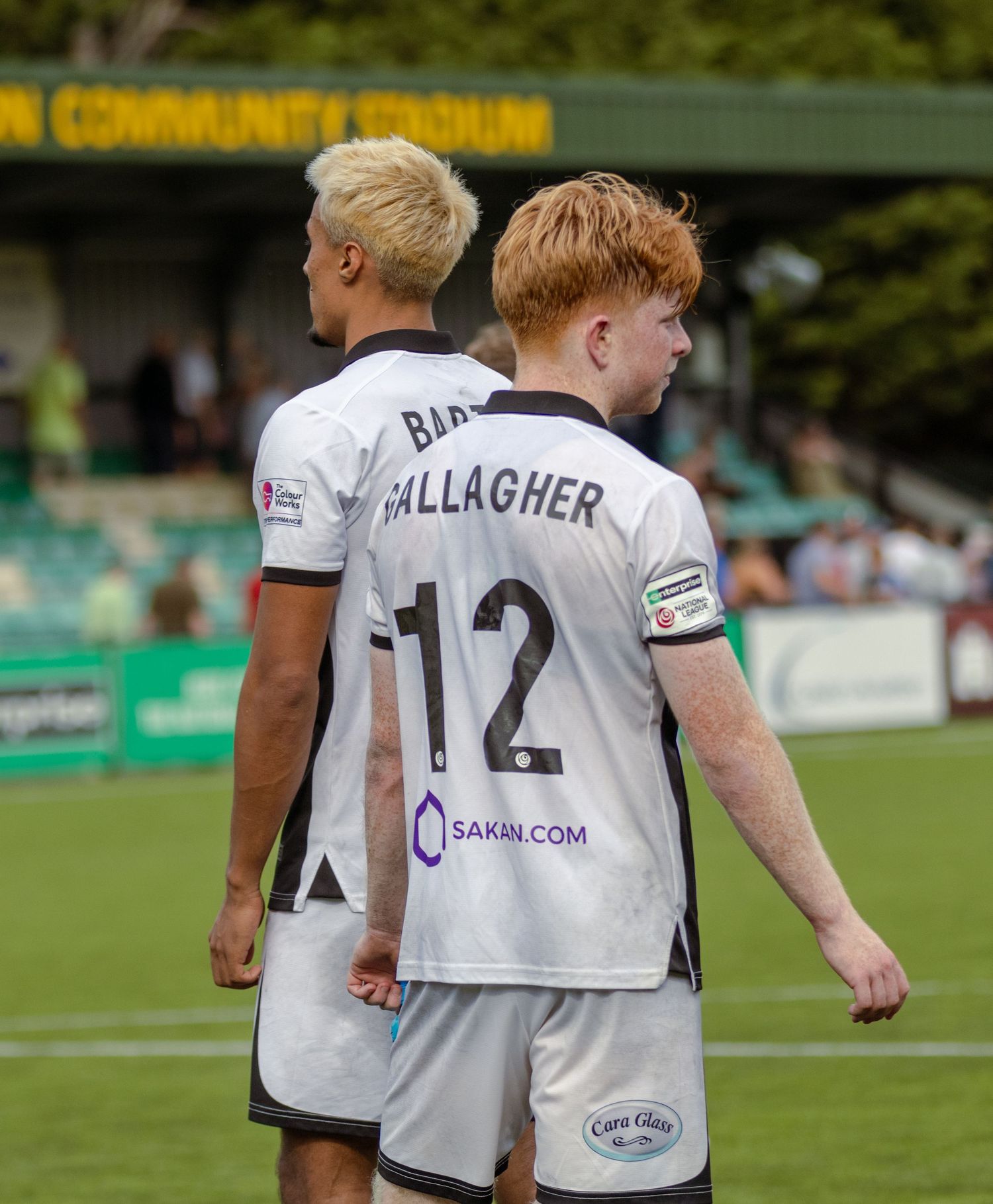 Two footballers in white away kits, including number 12 GALLAGHER, stand on a football pitch.