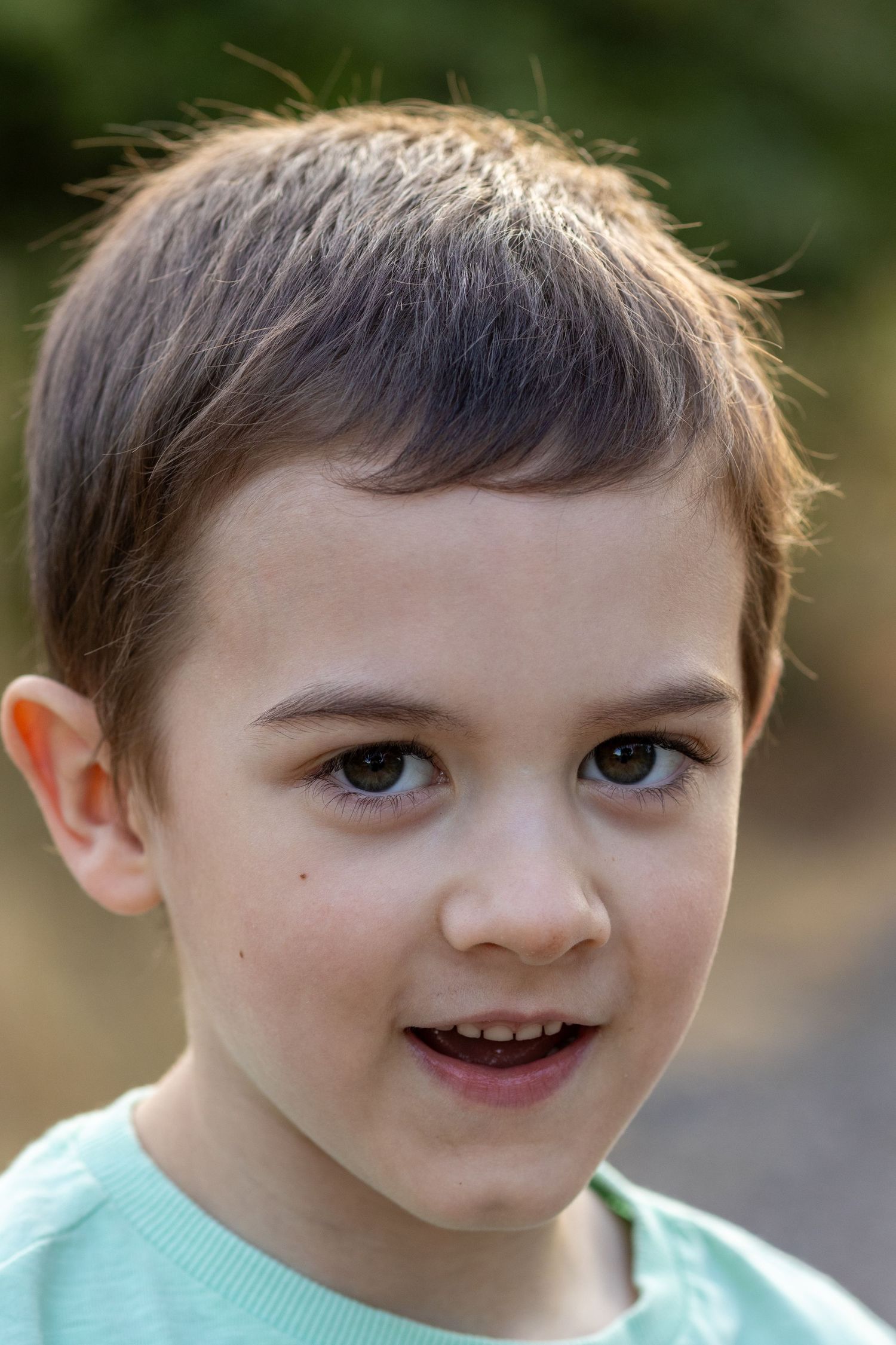 Smiling young boy with brown eyes and short hair in a green shirt photographed outdoors.