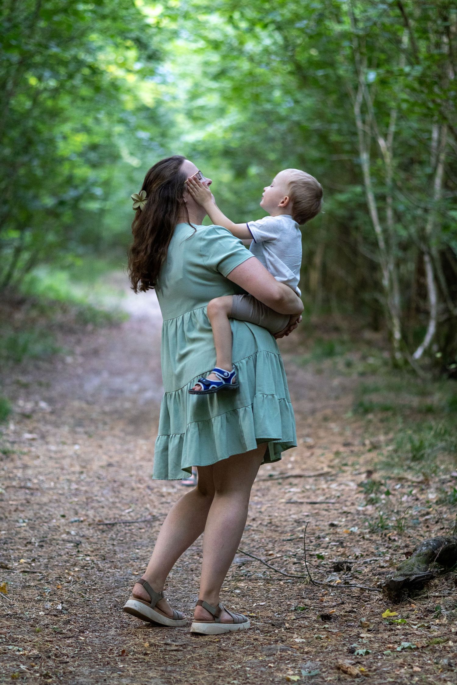 Woman in sage green dress carrying toddler on a leafy forest trail.