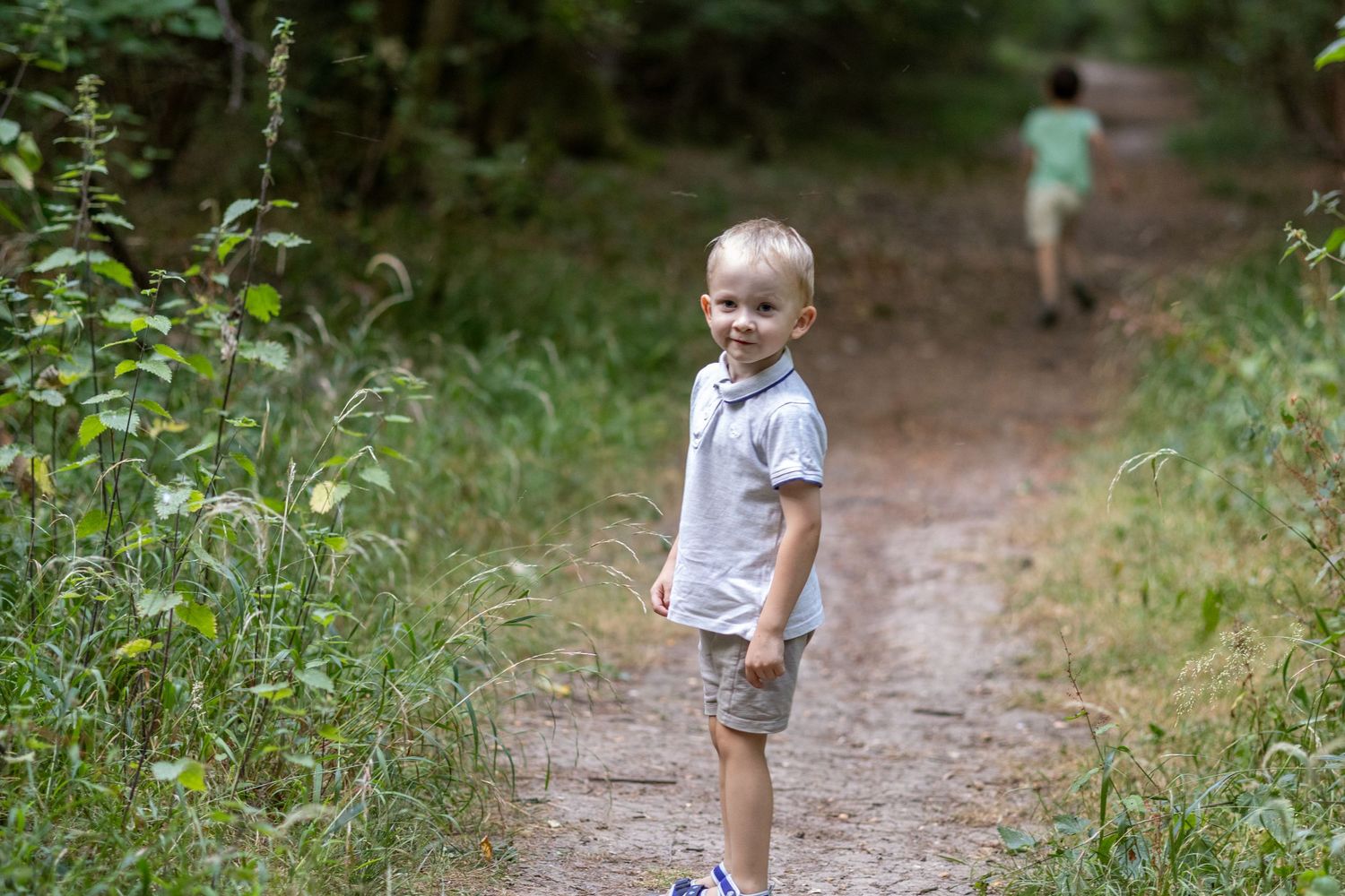 A smiling blonde toddler boy stands on a forest path with another child running behind.