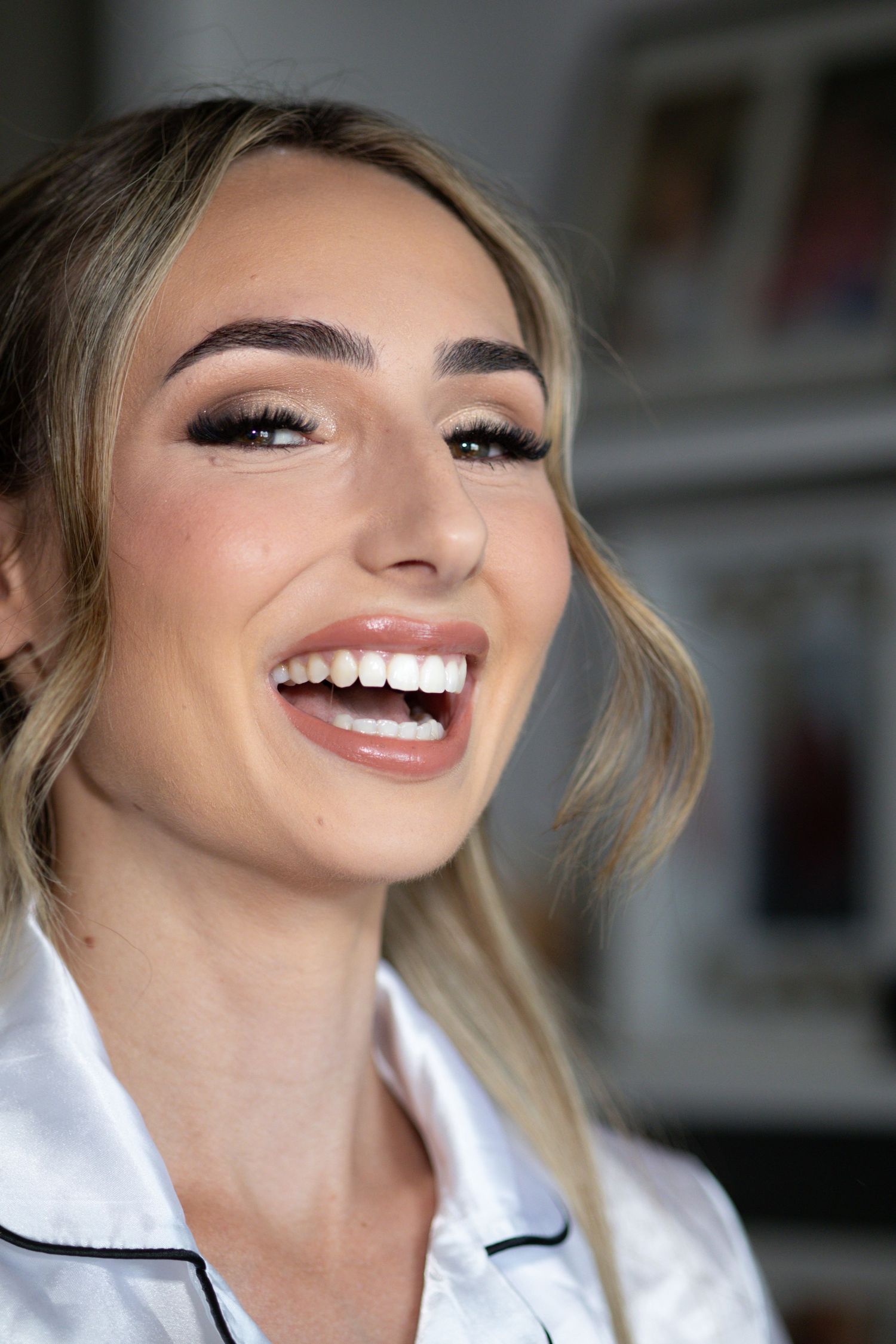 Joyful blonde woman laughing with glamorous bridal makeup and white satin robe in natural light.