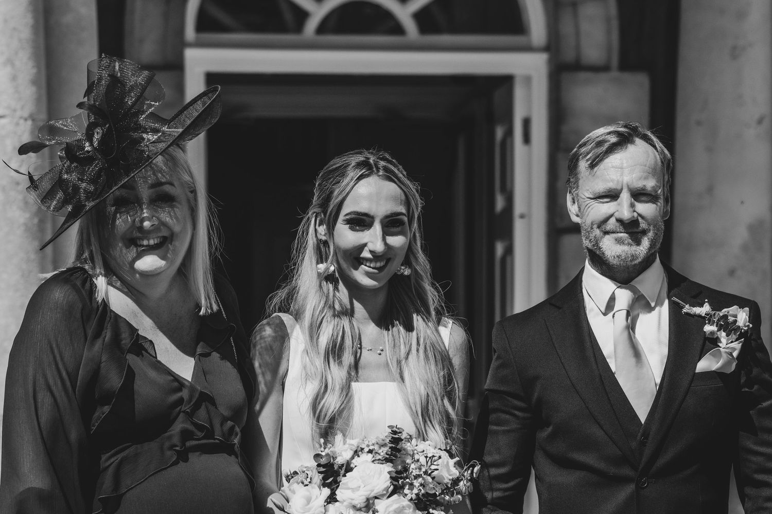 Black and white photo of bride with bouquet flanked by smiling woman in fascinator hat and man in suit at wedding.