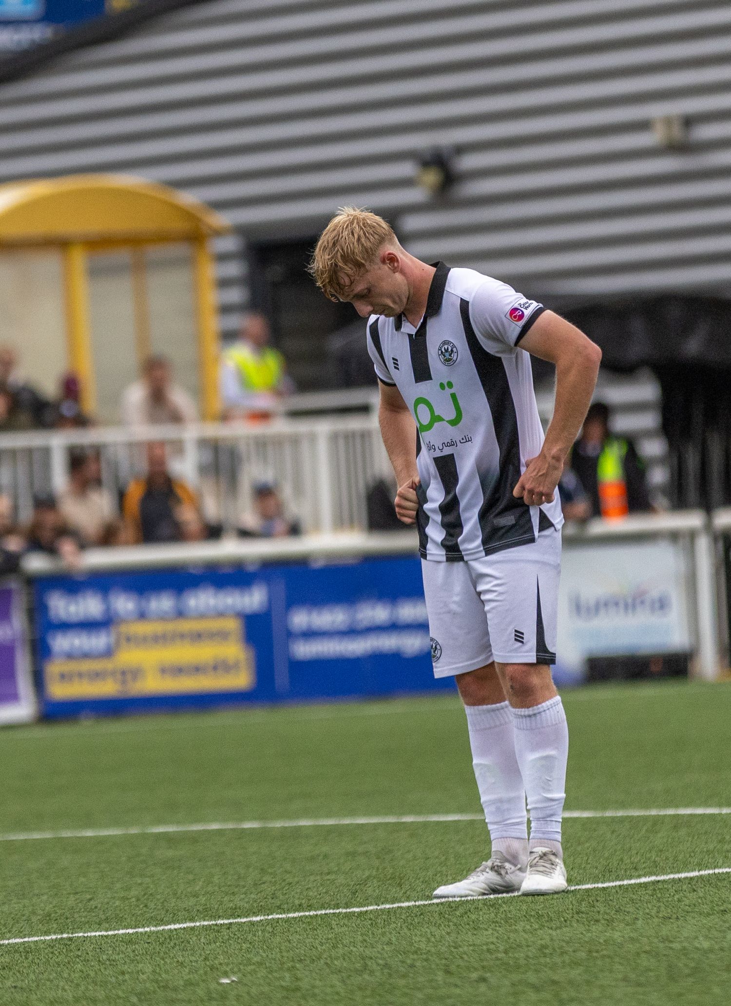 A young blonde footballer in white and black kit stands with hands on hips during a match at a non-league ground.