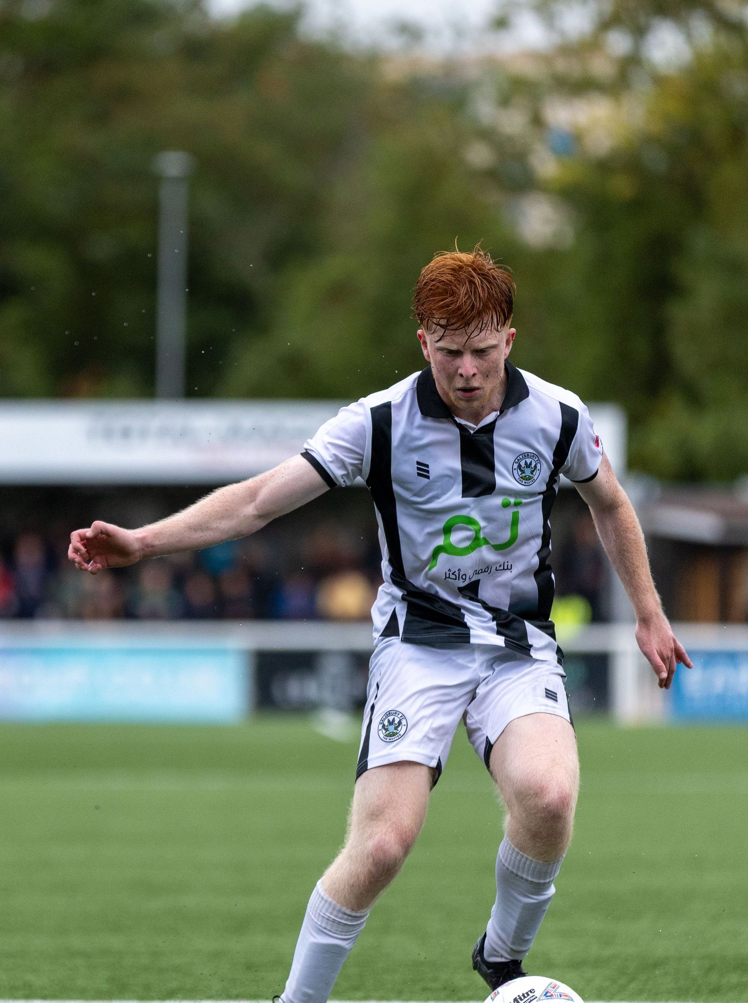 Young red-haired footballer in black and white kit dribbling a ball on a green pitch during a match.