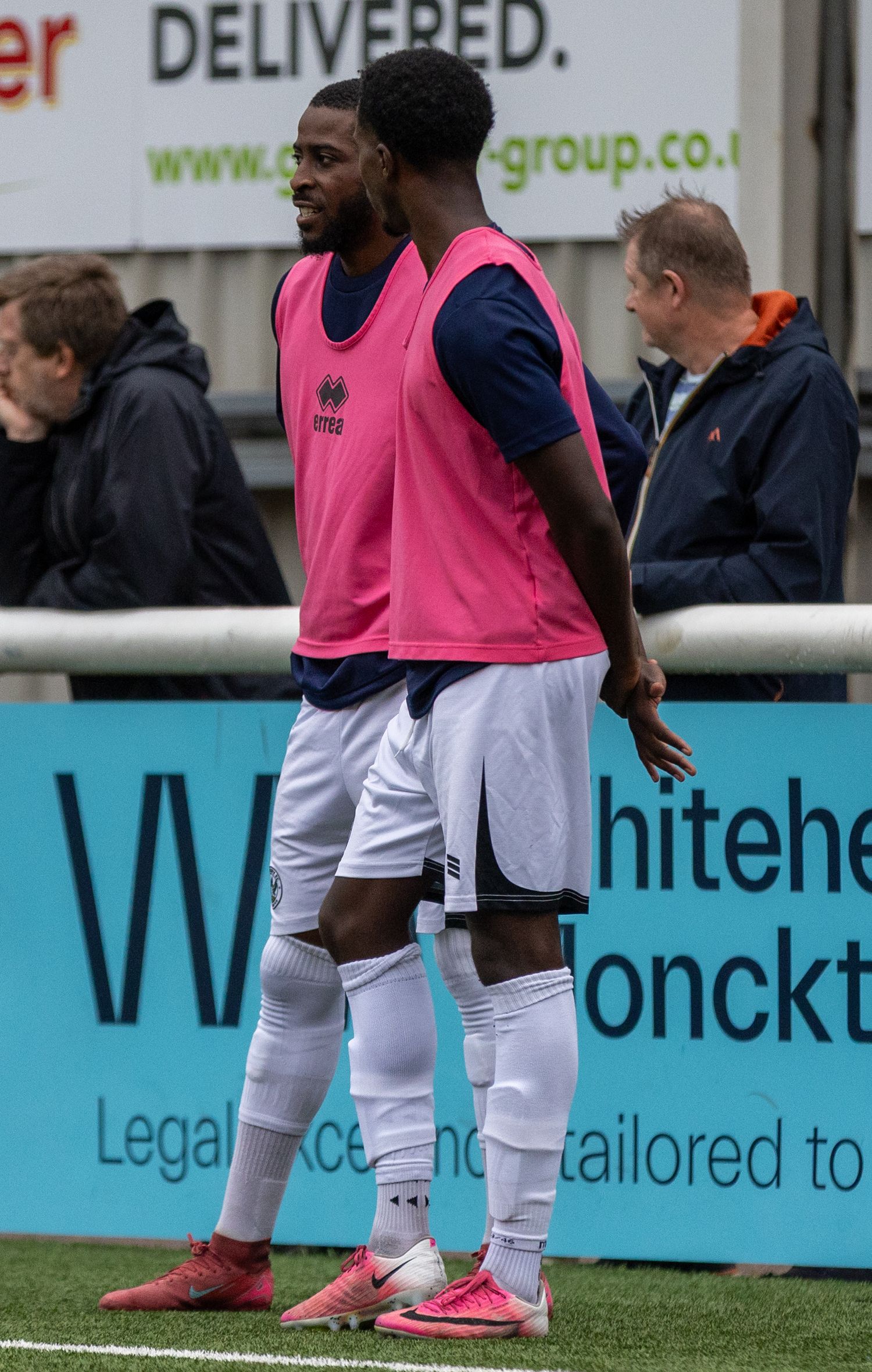Two football players in pink training bibs and white shorts stand on the sideline during a match.