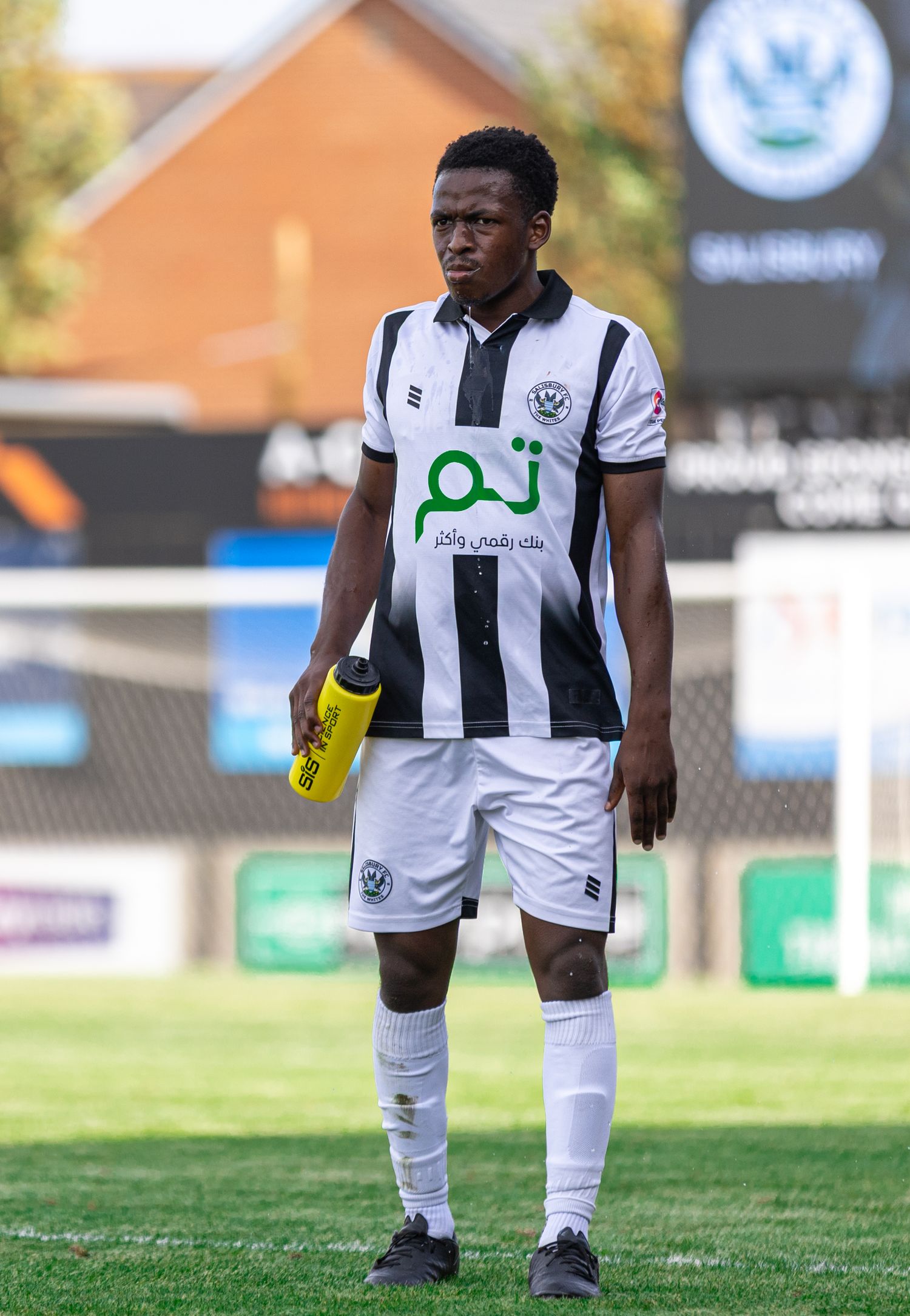 A football player in black and white striped kit holds a water bottle on a grass pitch during a match.