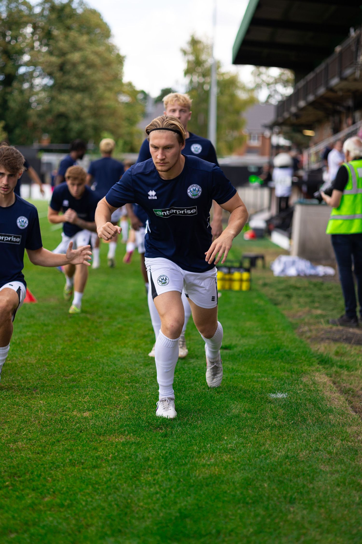 Soccer players in navy blue kits jogging onto a grass pitch at a non-league football ground on a cloudy day.
