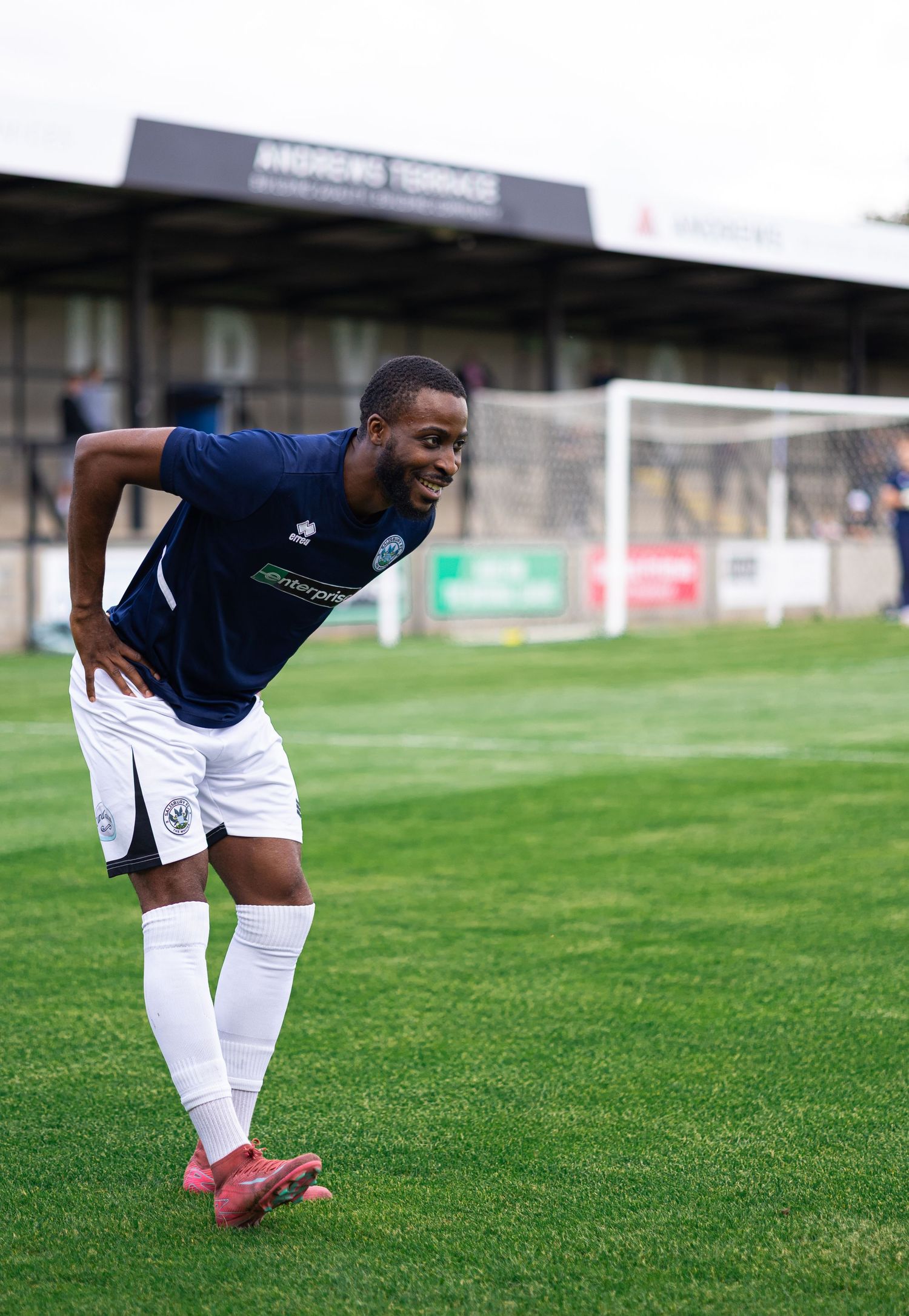 A soccer player in a navy blue kit smiles on a green pitch near the goal during a match.