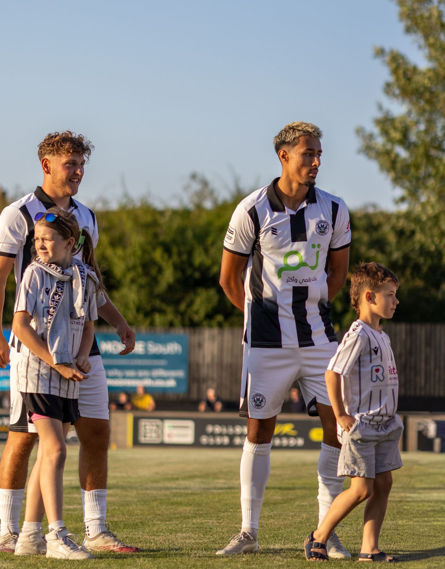 Two footballers in black and white kits walk onto the pitch accompanied by young mascots before kickoff.