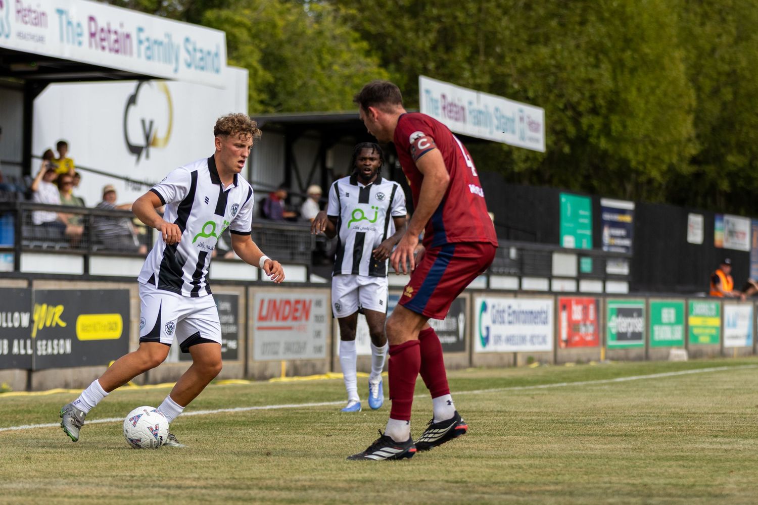 Two footballers challenge for the ball during a match, wearing black and white and red kits on a sunny grass pitch.