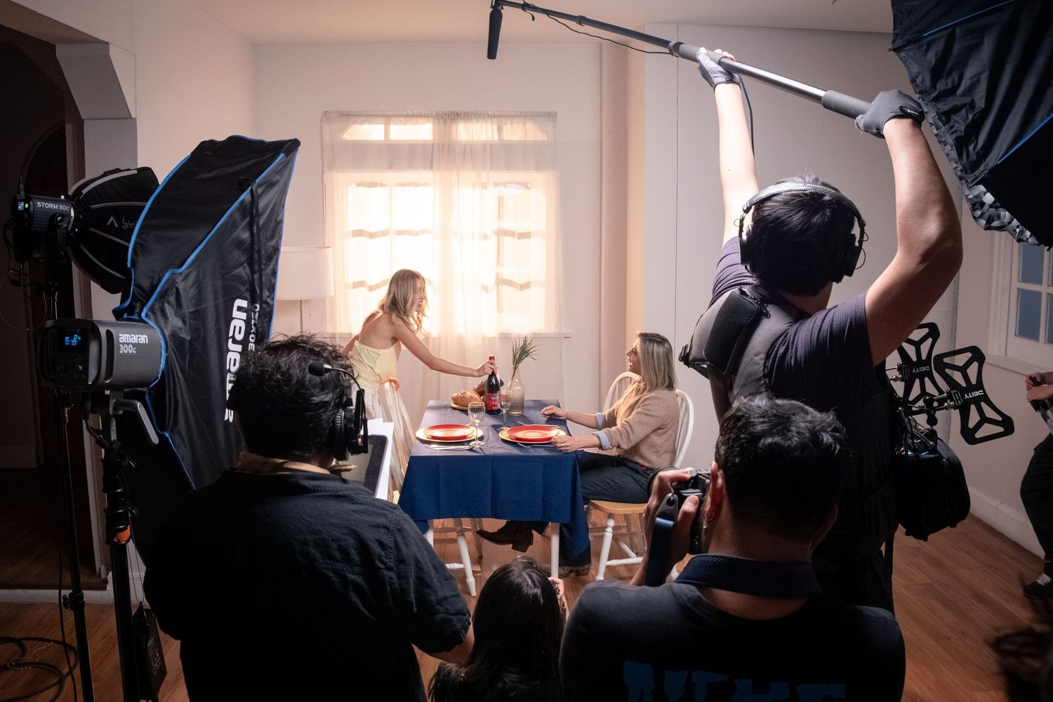 Film crew with boom mic and cameras shoots two women at a dinner table on a lit set.