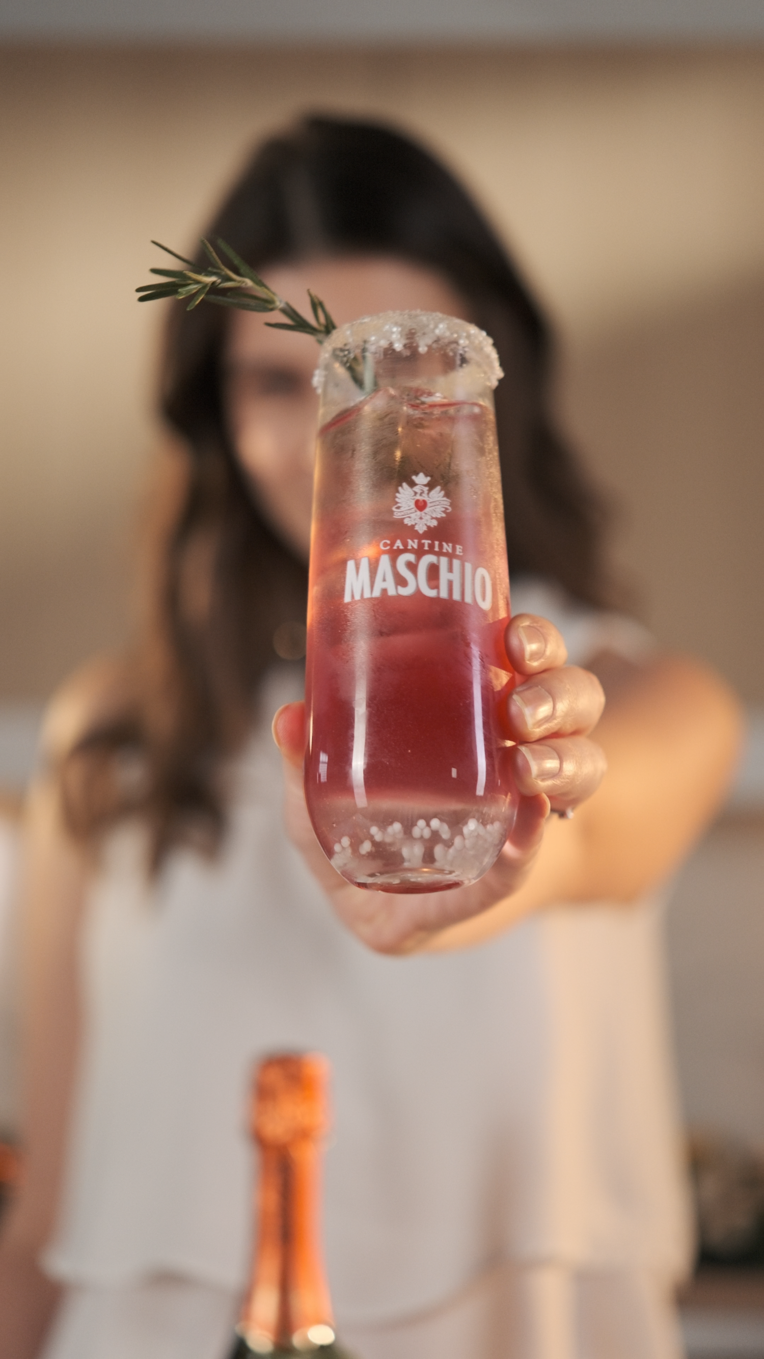 Woman holds Cantine Maschio branded cocktail glass with rosemary garnish toward camera.