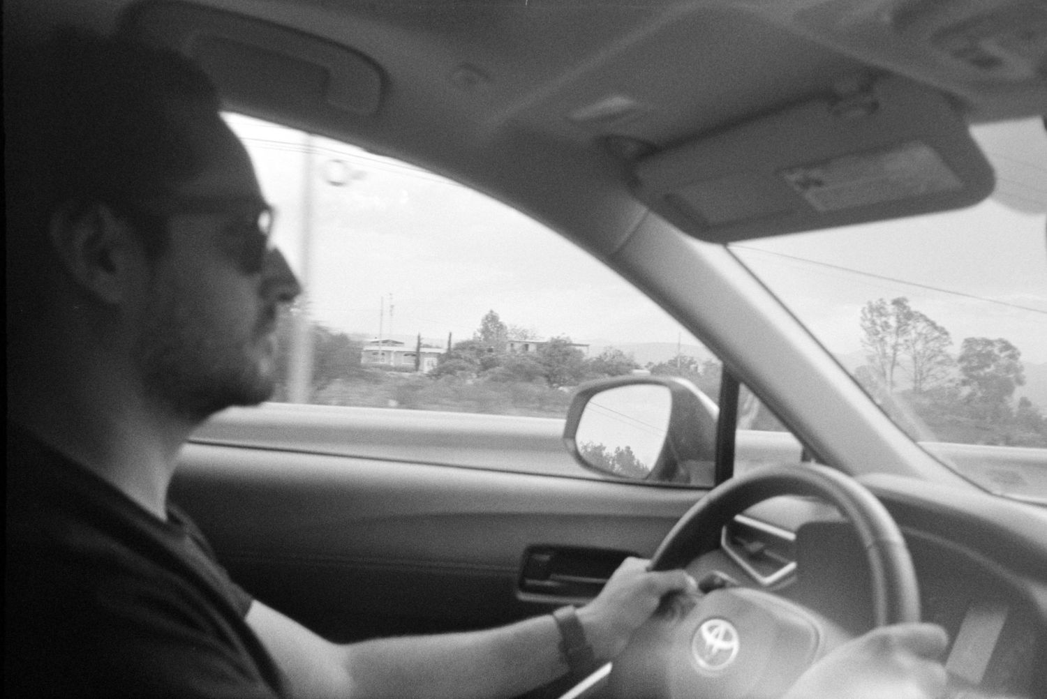 Black and white photo of bearded man driving car on highway wearing sunglasses.