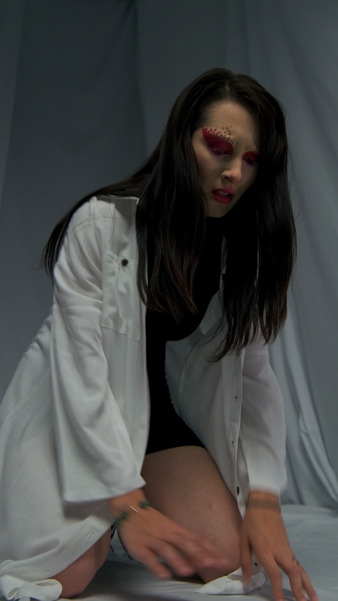 Dark-haired woman in white shirt with red eye makeup crouches dramatically on a white floor.