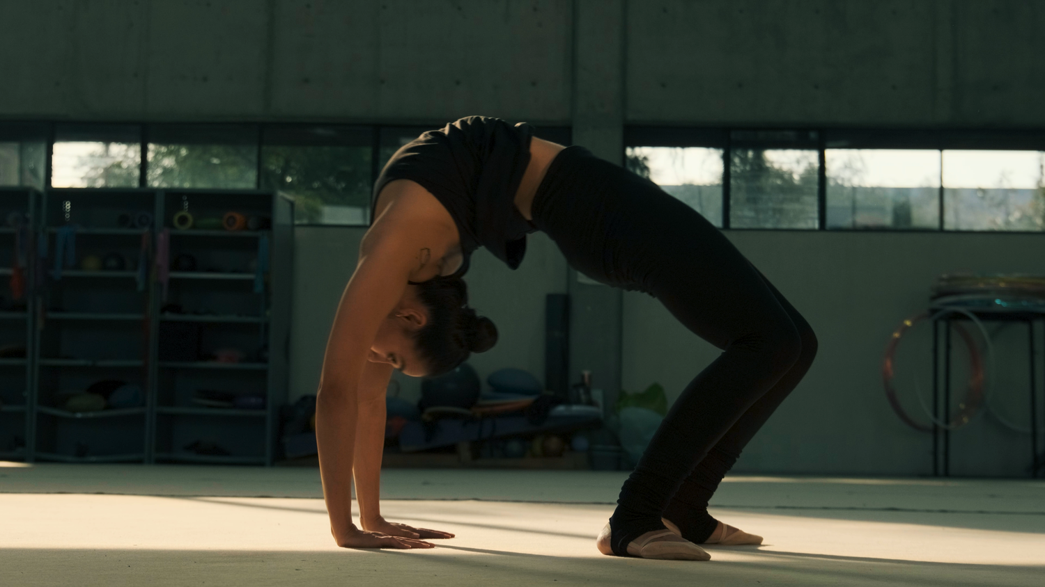A female gymnast performs a backbend stretch on the floor of a professional training gymnasium.