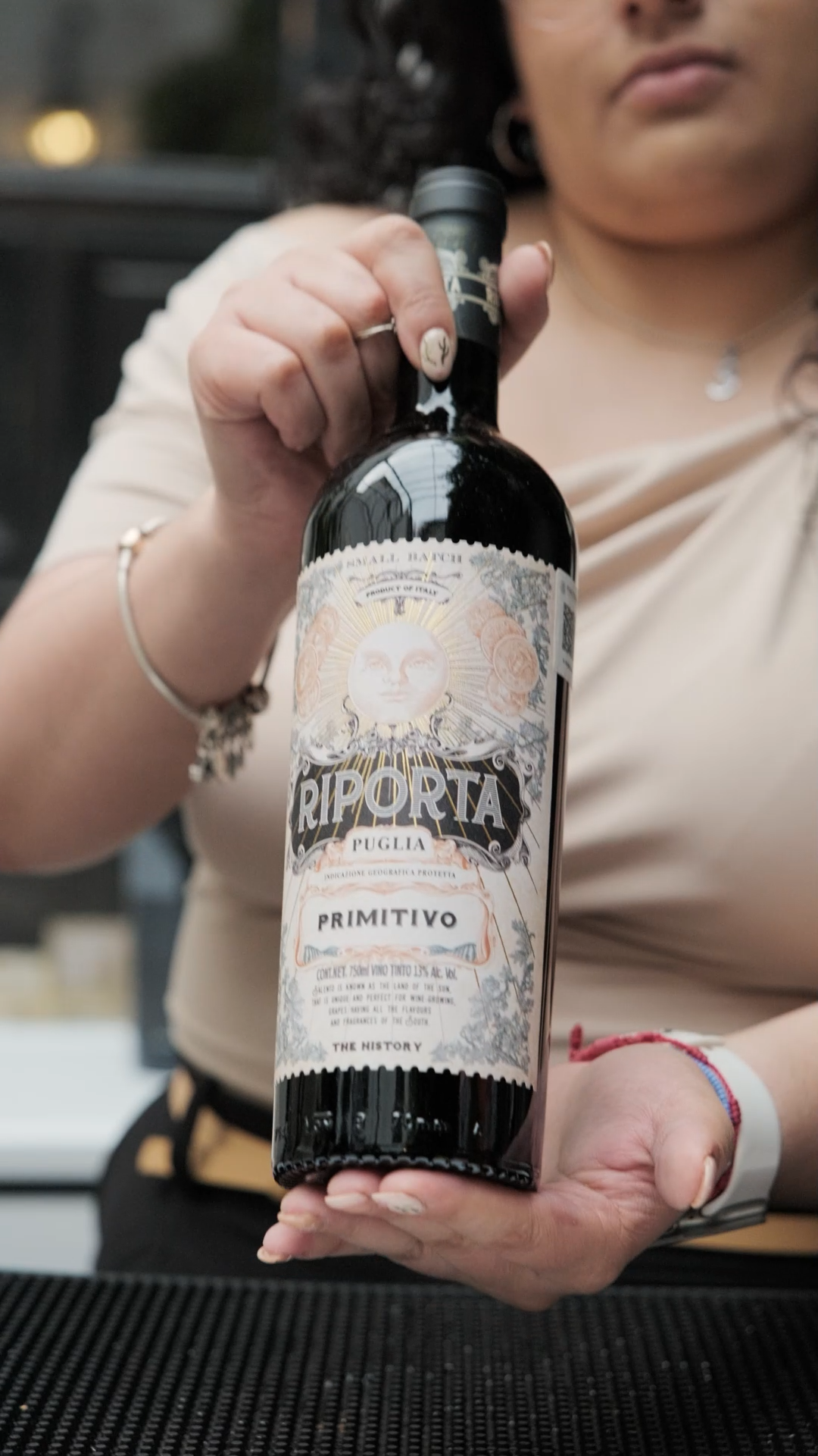 Woman holding Riporta Puglia Primitivo Italian red wine bottle with ornate vintage label design.