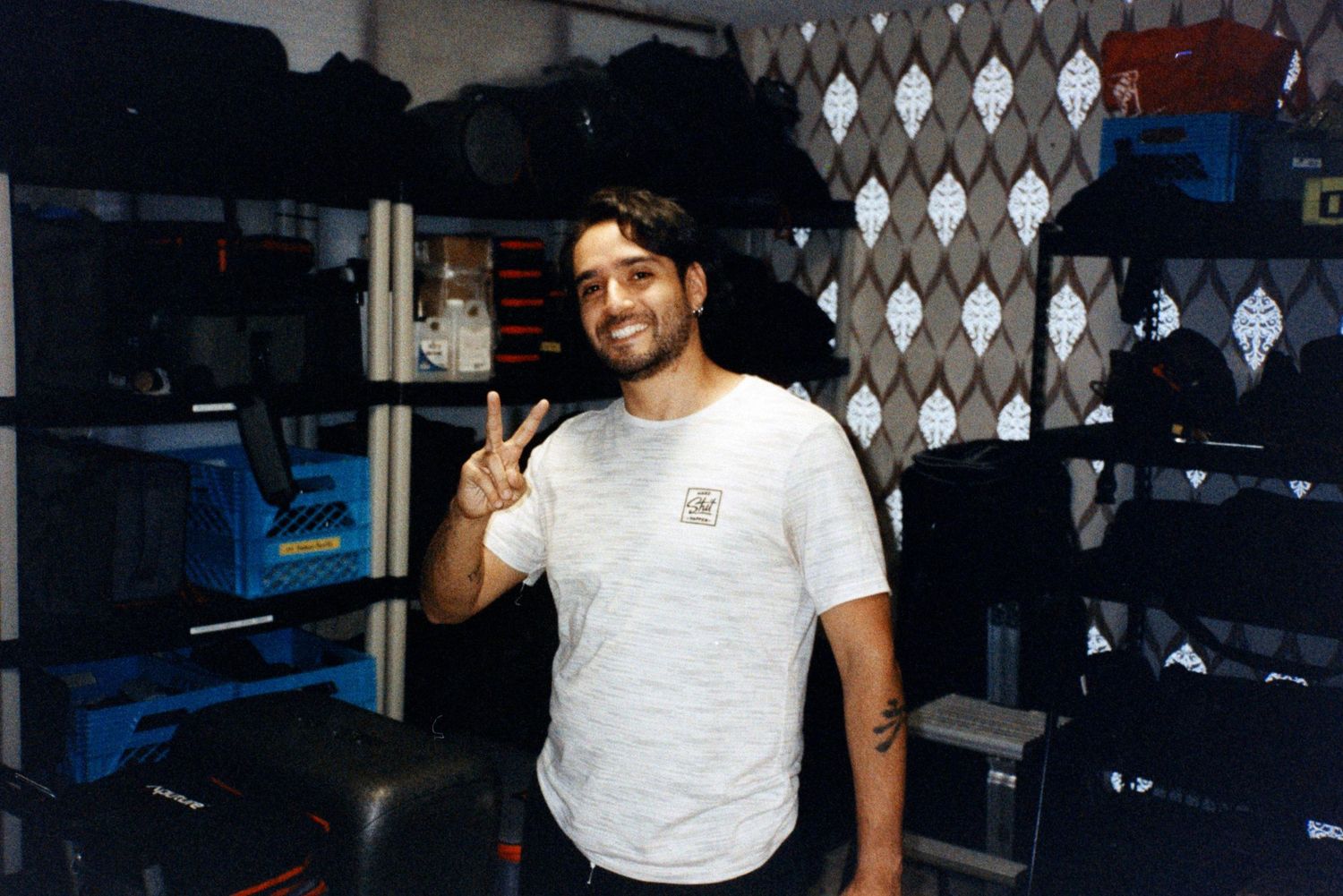 Smiling man in white t-shirt gives peace sign inside a storage room with equipment cases.