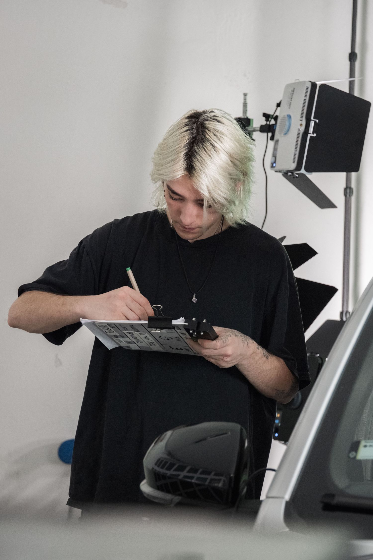 Blonde-haired crew member writes notes on a clipboard beside a car on a film set.