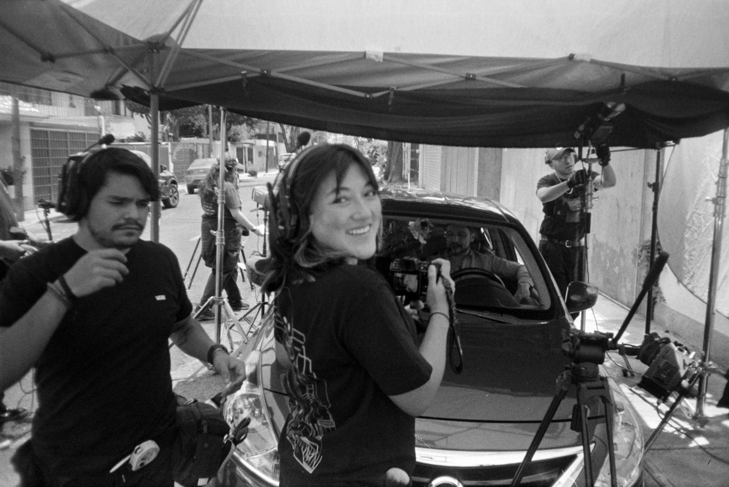 Black and white behind-the-scenes film set with crew members smiling near camera equipment and a car outdoors.