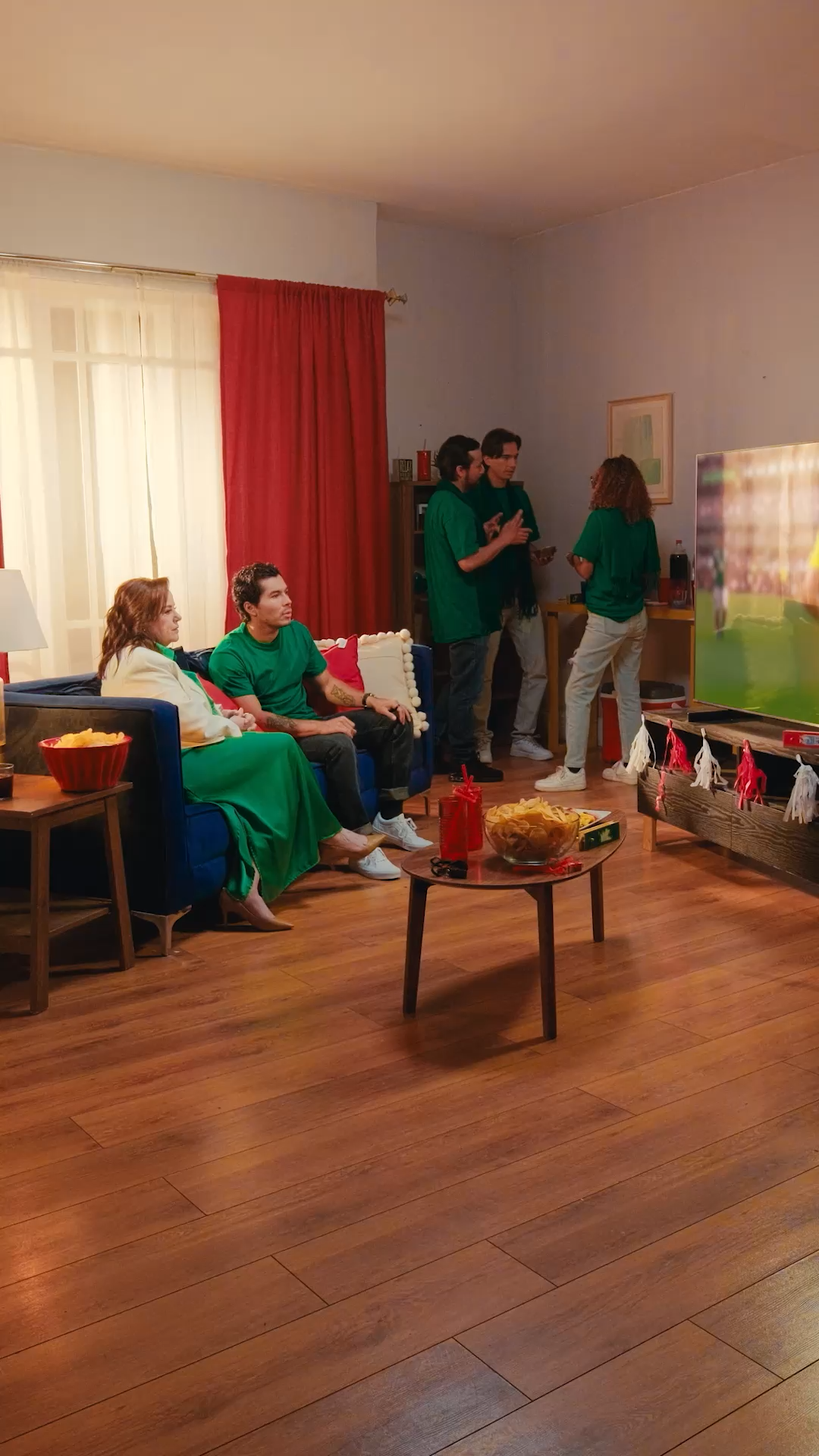 A group of friends in green shirts watch soccer on TV in a cozy living room with snacks.