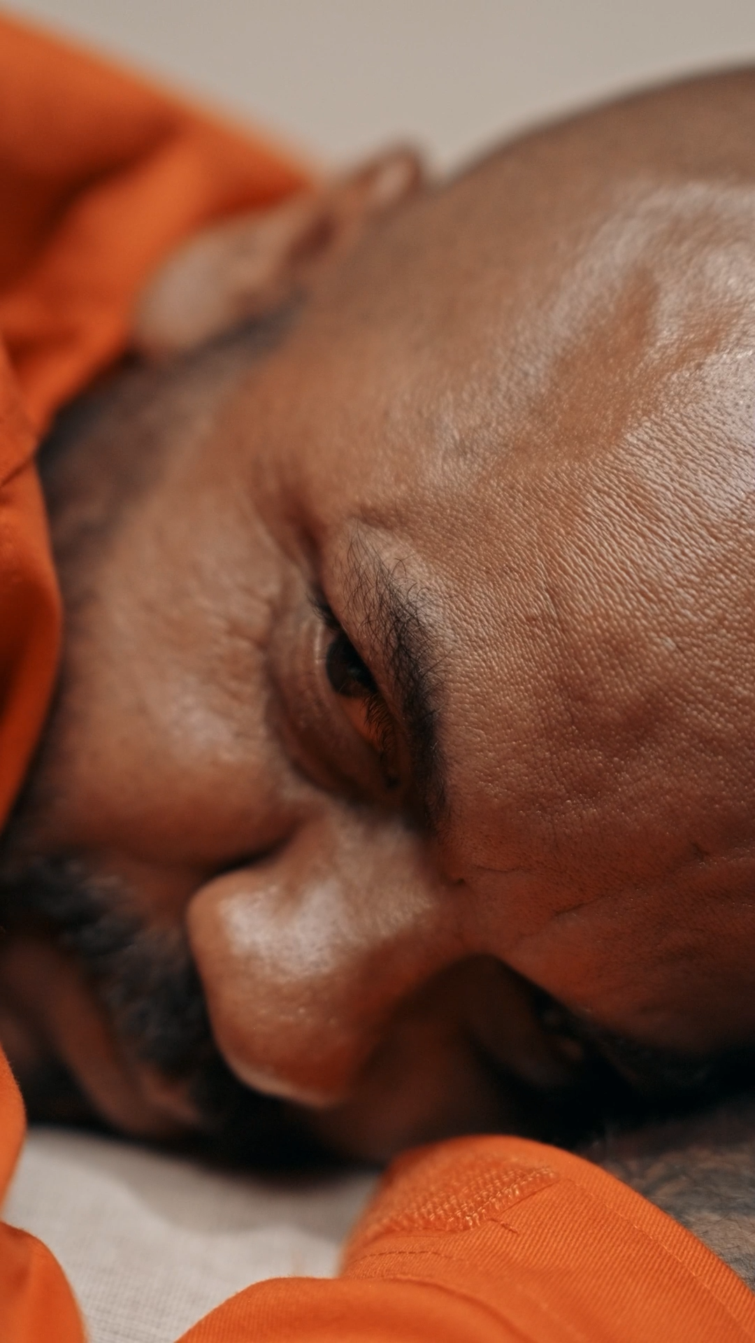 Close-up of a bald man in an orange jumpsuit lying down with eyes closed, resting.
