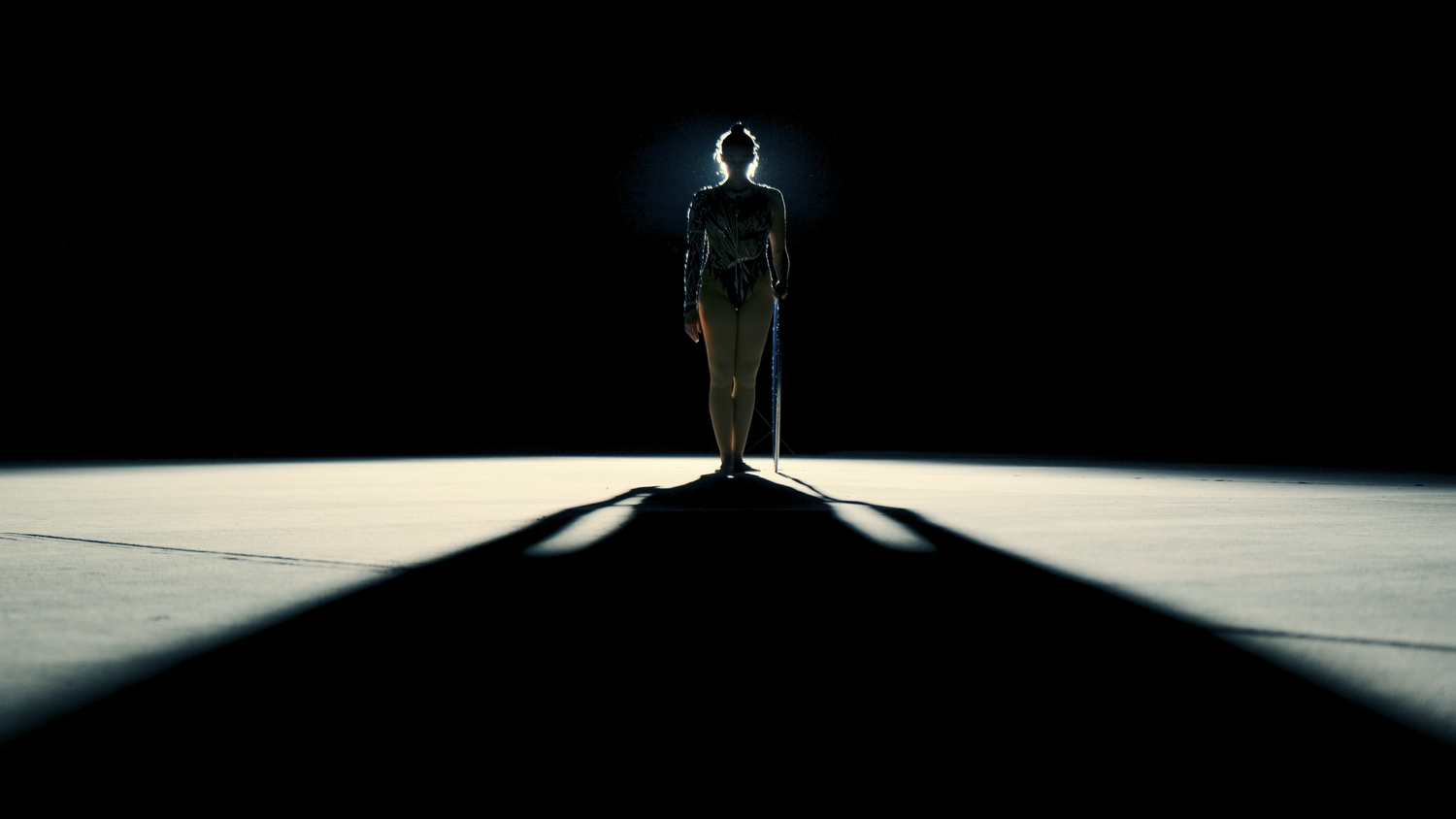 Silhouetted gymnast standing under spotlight casting dramatic shadow on arena floor.