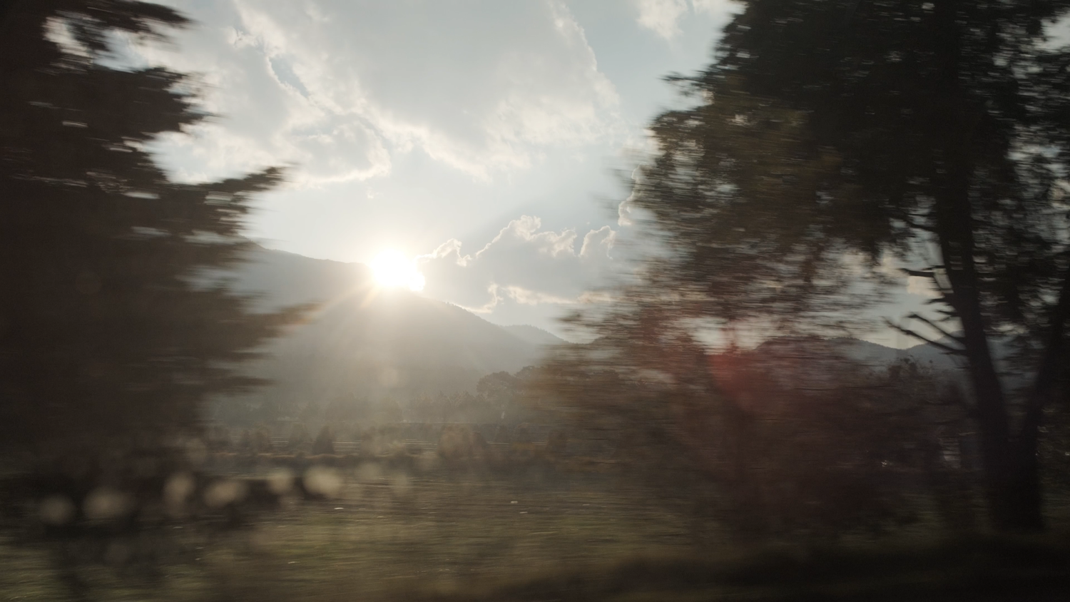 Scenic view of sunlight breaking through trees over misty mountains captured through a rain-spotted window.