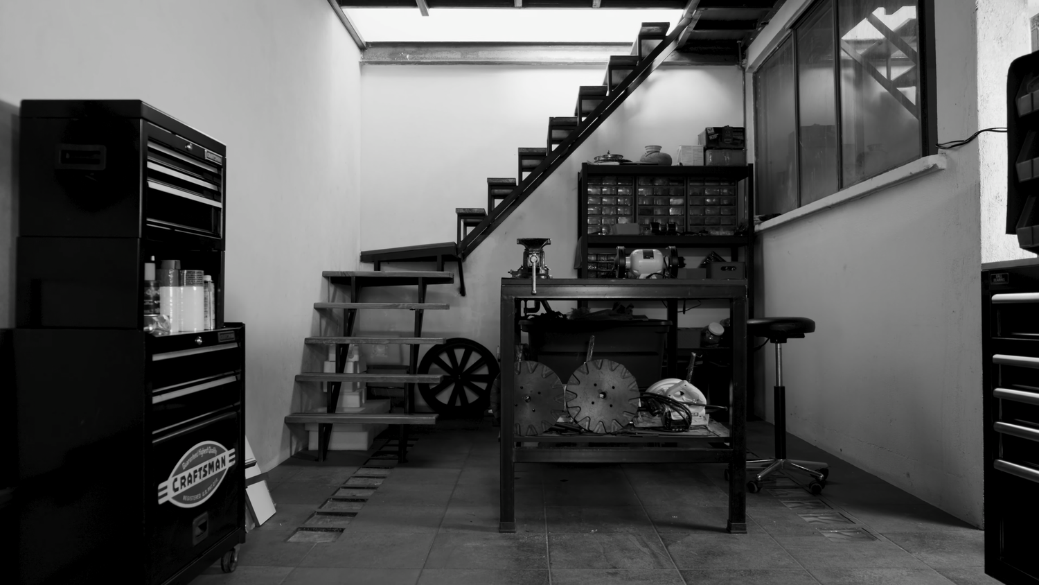 Black and white image of garage interior with stairs, tool chest, and metal shelving unit.