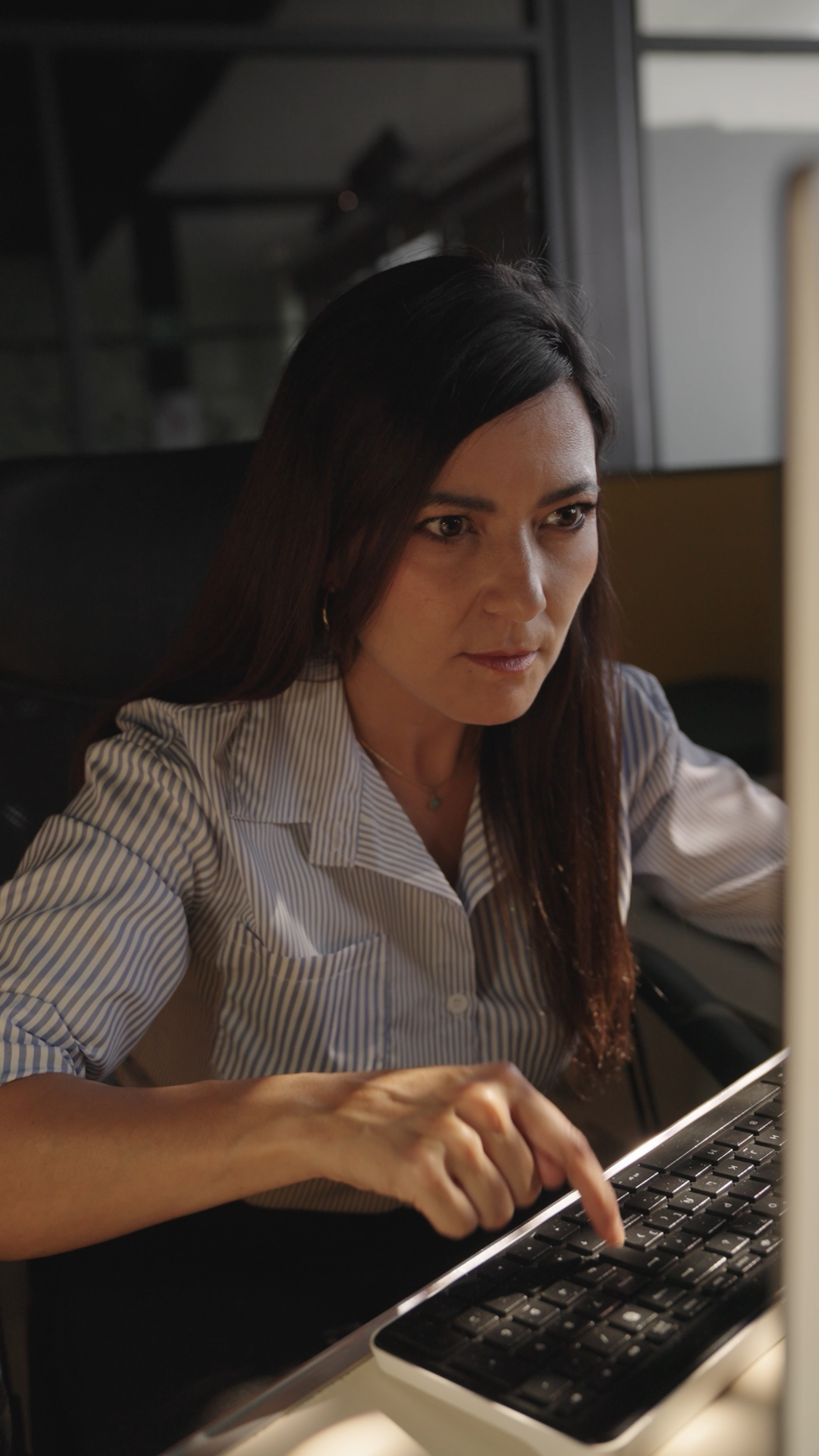 Focused woman with dark hair typing on a keyboard at a computer in a dimly lit office.