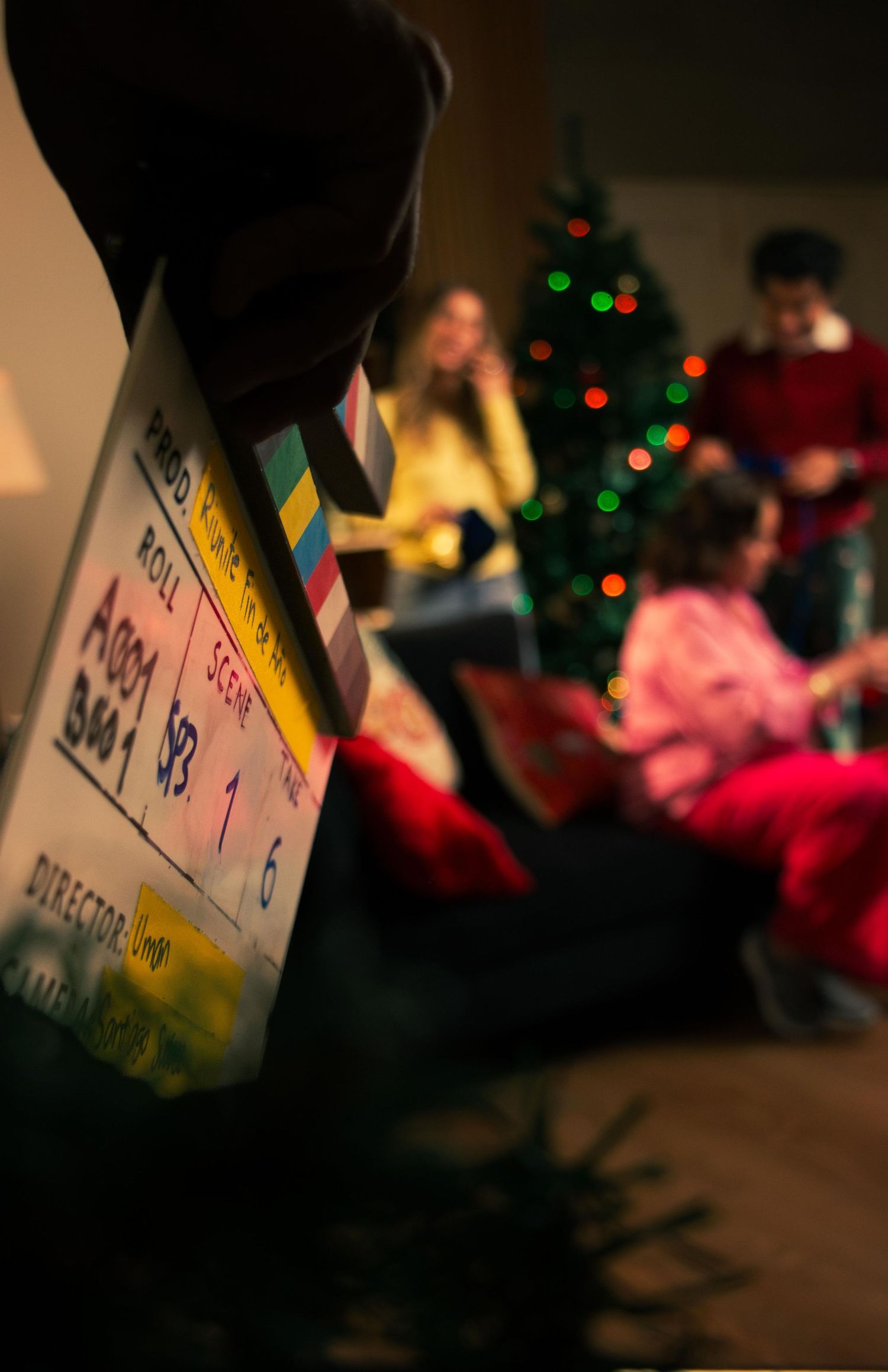 Close-up of a film clapperboard with a decorated Christmas tree and actors visible in the background.