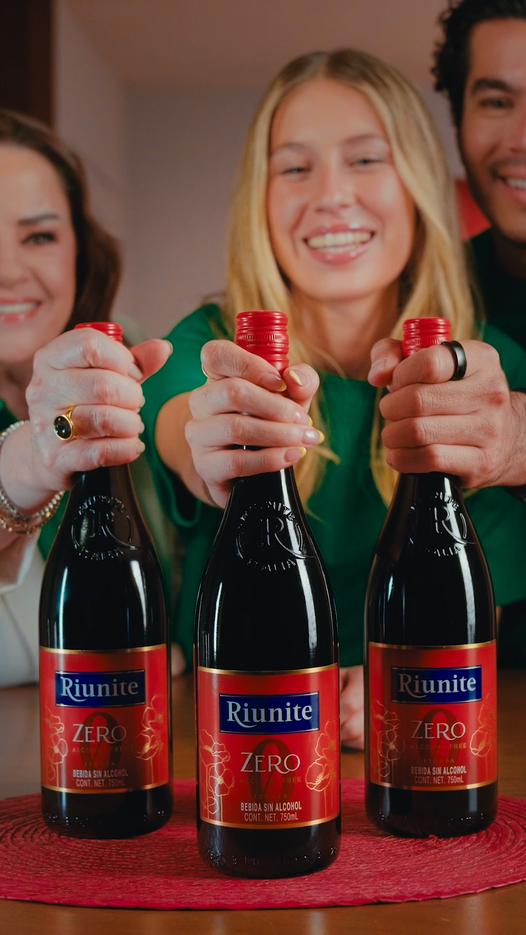 Three people hold Riunite Zero non-alcoholic beverage bottles together at a social gathering.